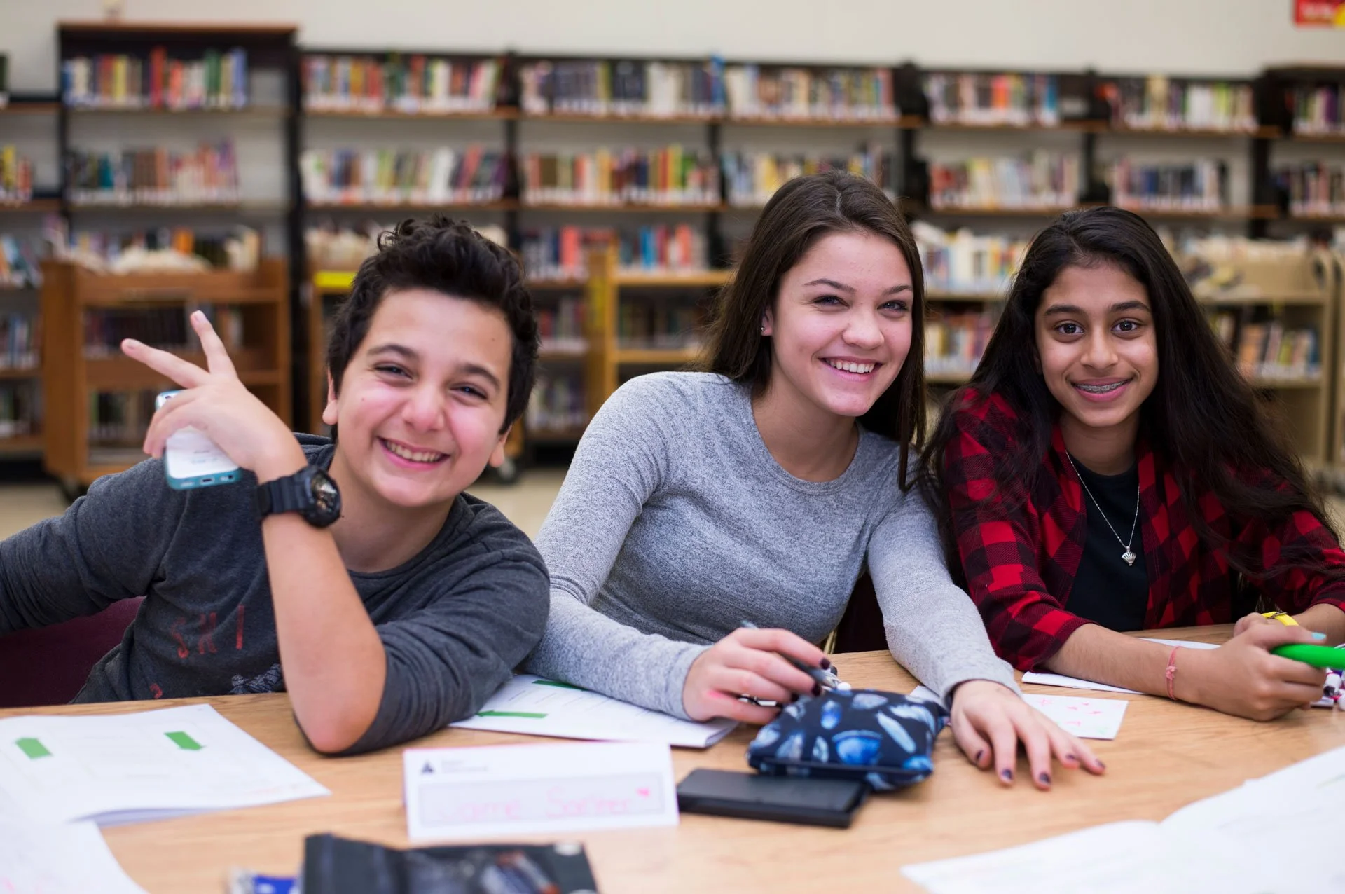 Three students are smiling at a table in a library during a JA (Junior Achievement) Southern Alberta program, holding pens and a remote.