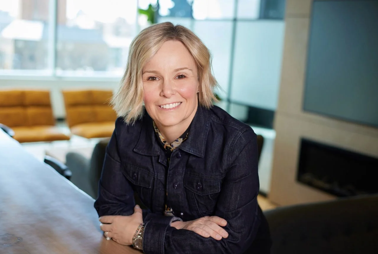 A woman with short blonde hair and a smile, sitting at a table in a modern office or conference room with large windows and a yellow sofa in the background.