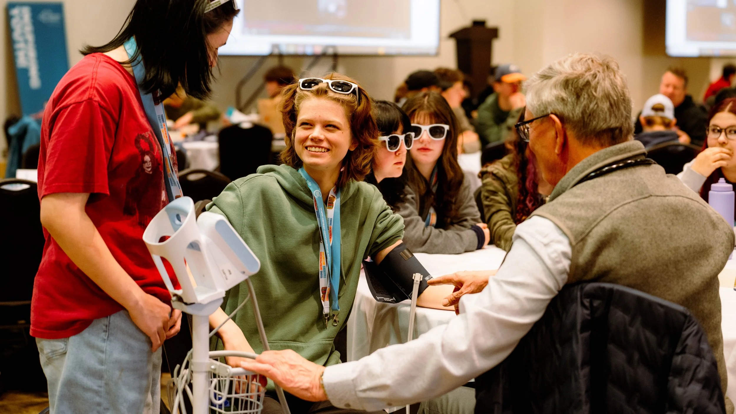 A young woman with a green hoodie smiling as she receives a health checkup from an older man with glasses, in a busy conference room with other people, some wearing sunglasses, in the background.