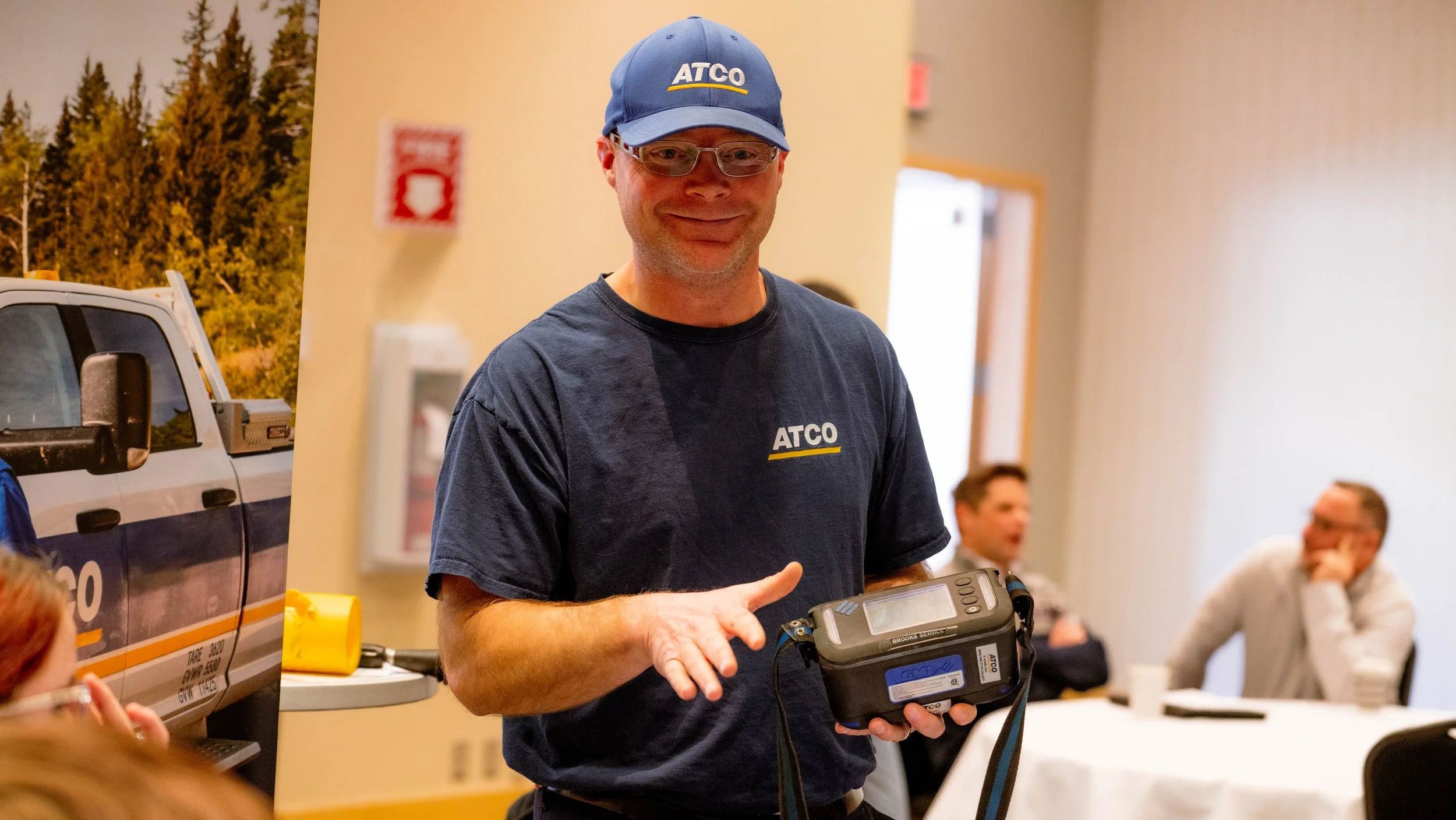 A man wearing glasses, a ATCO cap, and a navy T-shirt with the ATCO logo, holding a portable electronic device, inside a room with several people in the background.