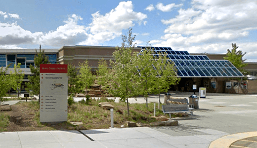 A modern building with solar panels on the roof, surrounded by trees, benches, and signage, with a partly cloudy sky overhead.