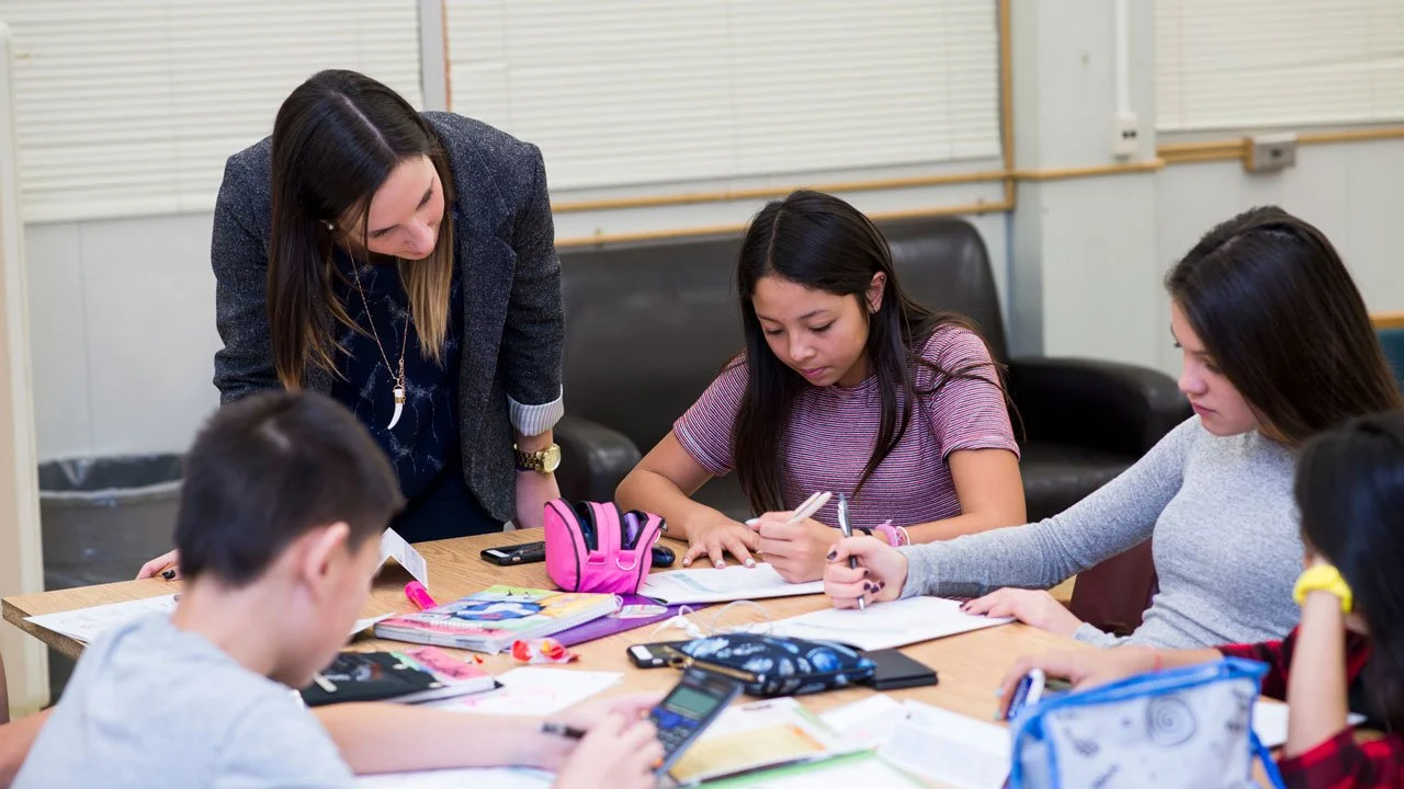 Teacher assisting students with homework at a table in a classroom.