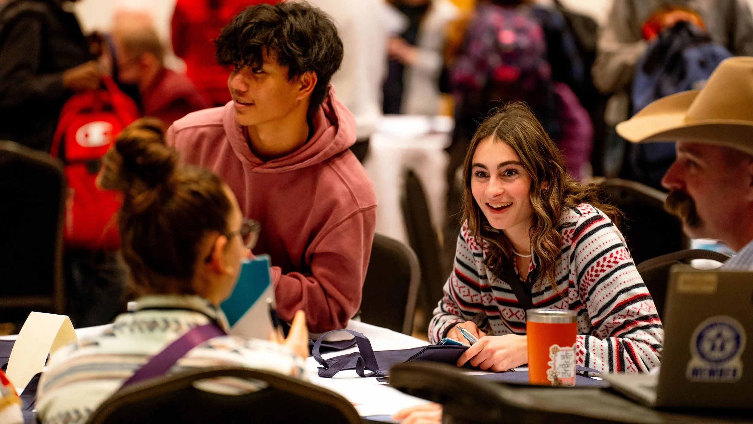 Young man in a pink hoodie and young woman in a festive sweater smiling and talking at a table surrounded by people at a social gathering.