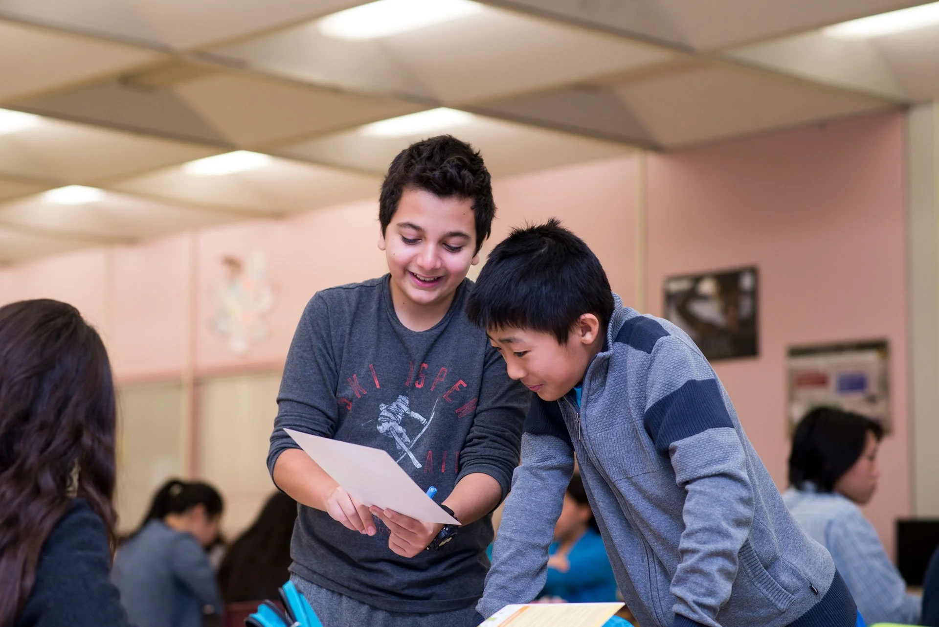 Two boys, one with curly dark hair and the other with straight black hair, are smiling and leaning over a table, looking at a piece of paper together at a classroom or library. The background shows other children sitting and a pink wall.
