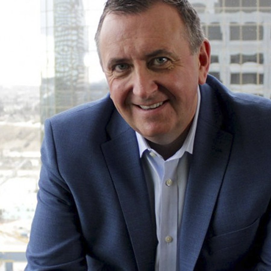 A man in a blue suit smiling in front of a large window with a cityscape background. Kent MacDonald, Vice Chair

Director
