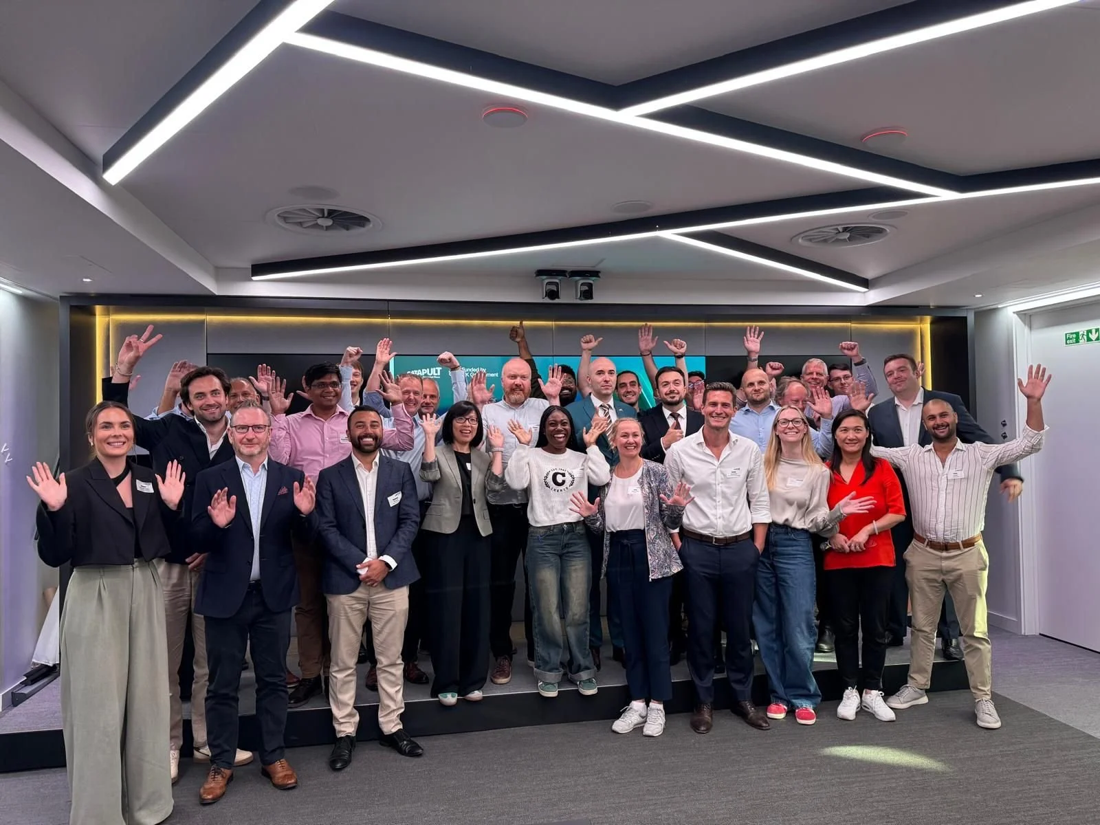 Group of diverse professionals smiling and waving in a modern conference room during a corporate event or meeting.