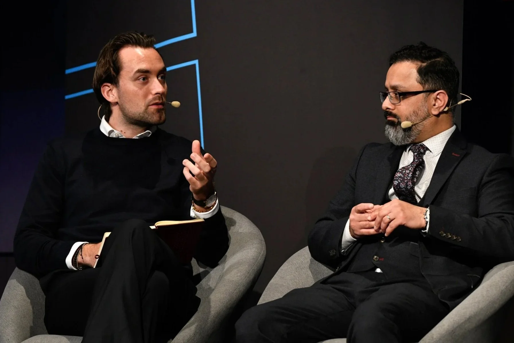 Two men sitting on gray chairs engaged in conversation, wearing microphones, dark background.