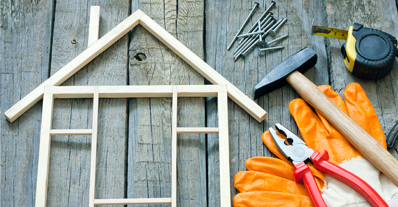 Tools and materials for building a small wooden house, including wooden frames, nails, hammer, tape measure, wire cutters, and gloves on a wooden surface.