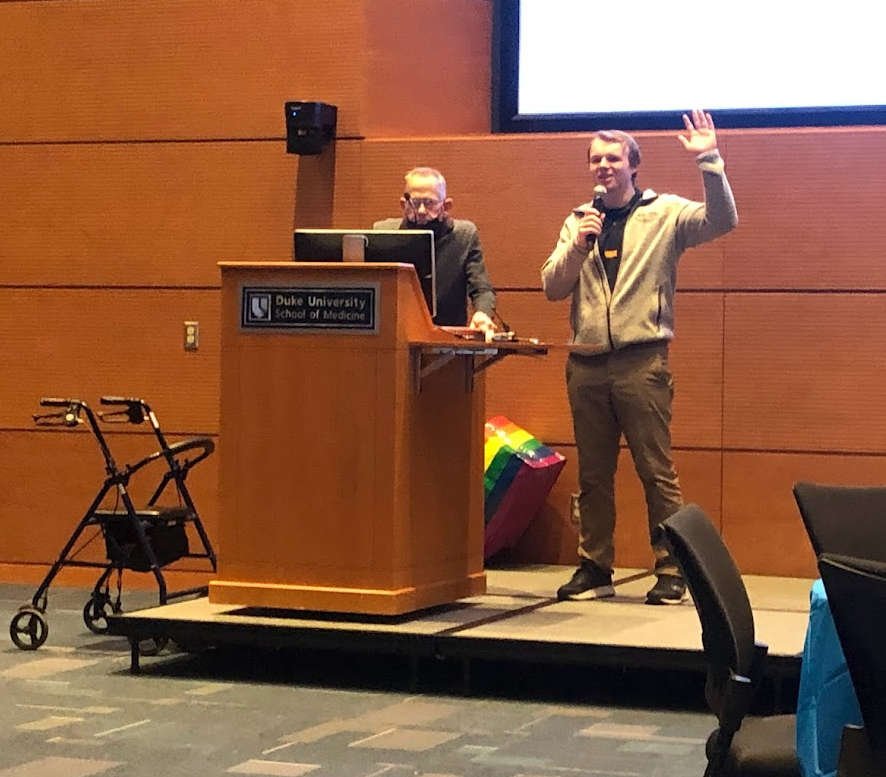 Two men speaking at a podium in a conference room at Duke University School of Medicine, with a large screen behind them. Presenting on aging at home safely and duke hotspotting initiative.