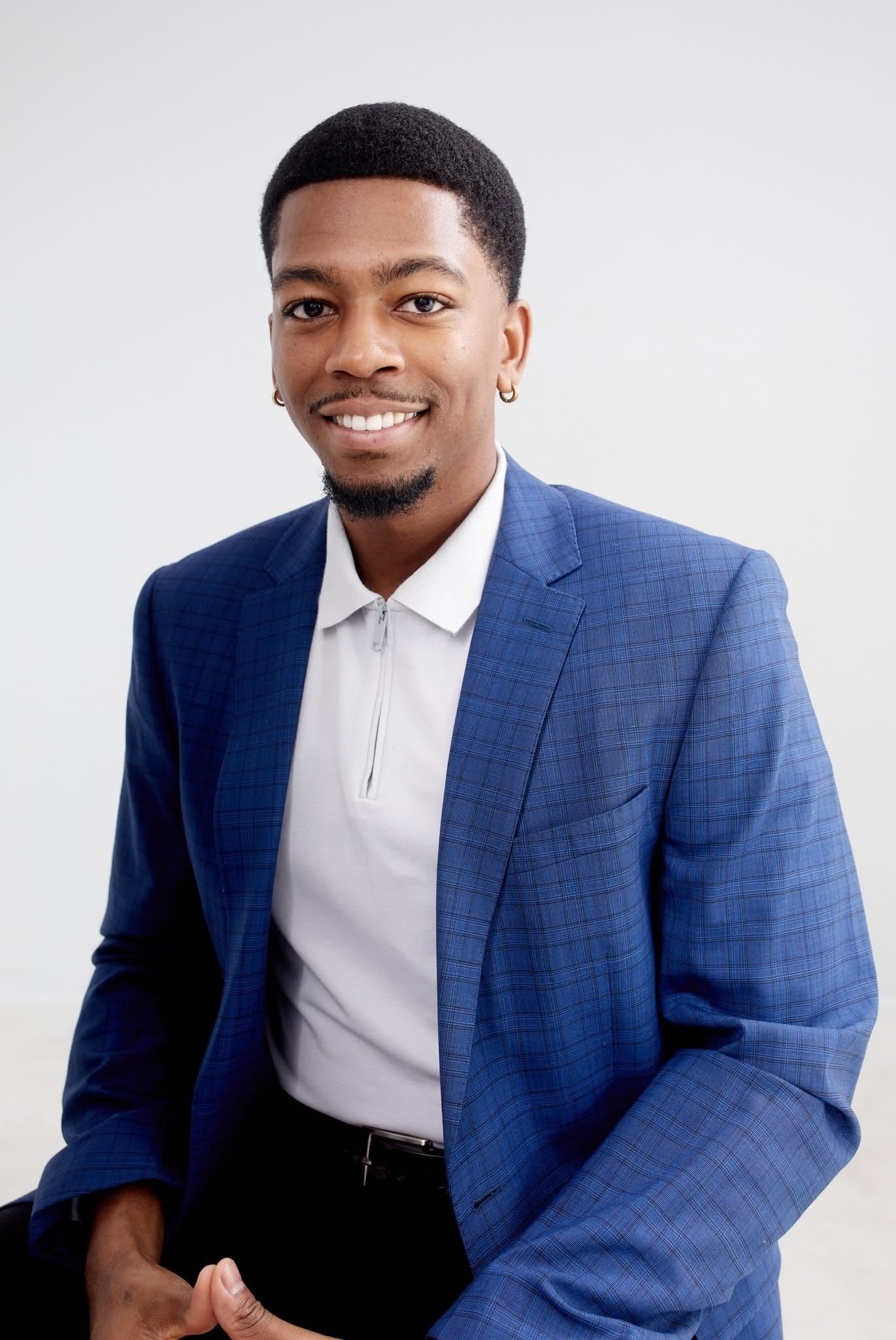 A portrait of a smiling Black man in a blue blazer and white zip-up shirt, sitting against a plain white background.