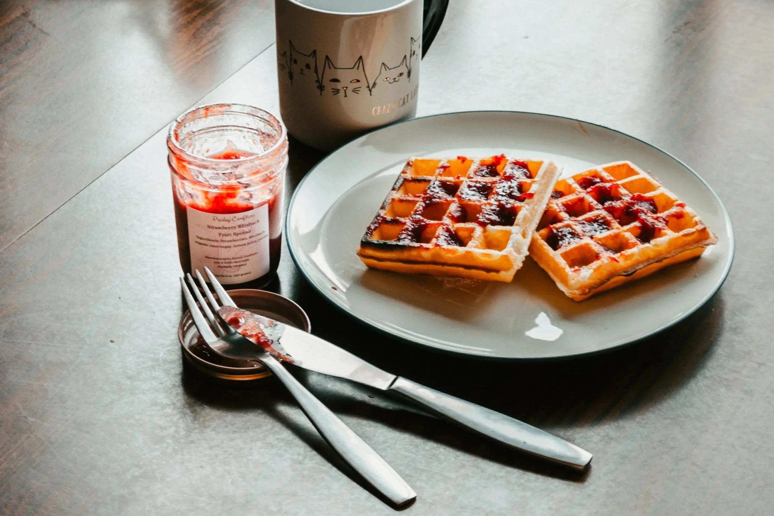 Morning waffles, coffee in a crazy cat mom mug with a jar of Strawberry Rhubarb Jam on the side