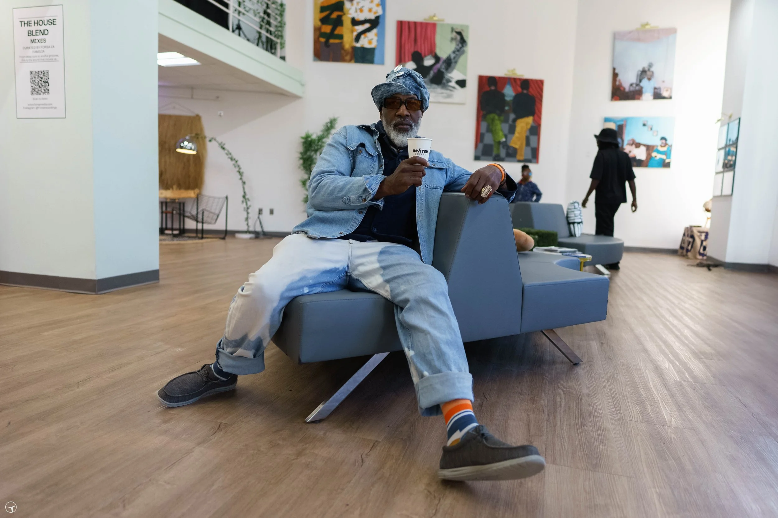 An older man with gray hair and a beard sitting on a light gray sofa in an art gallery, holding a paper cup labeled 'INVITED,' with colorful paintings on the walls behind him, and a woman in black walking away in the background.