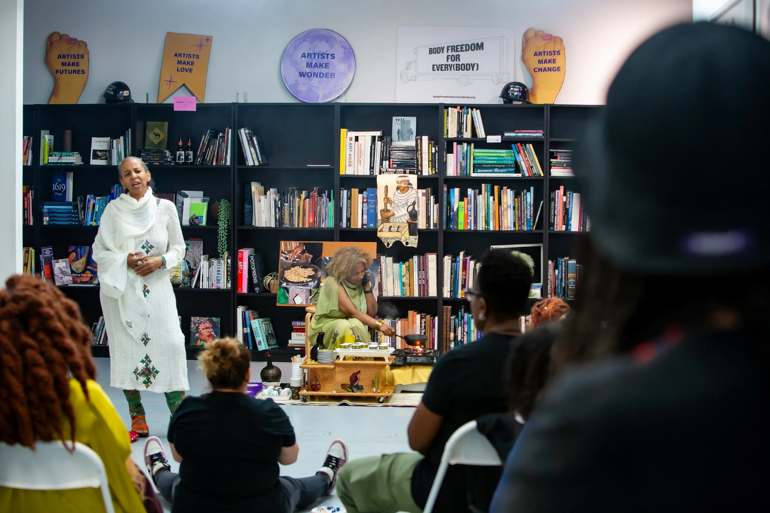 A woman dressed in white standing and speaking during a multicultural event, with seated audience and a woman in traditional attire preparing food on the floor in front of a bookshelf decorated with posters and signs.