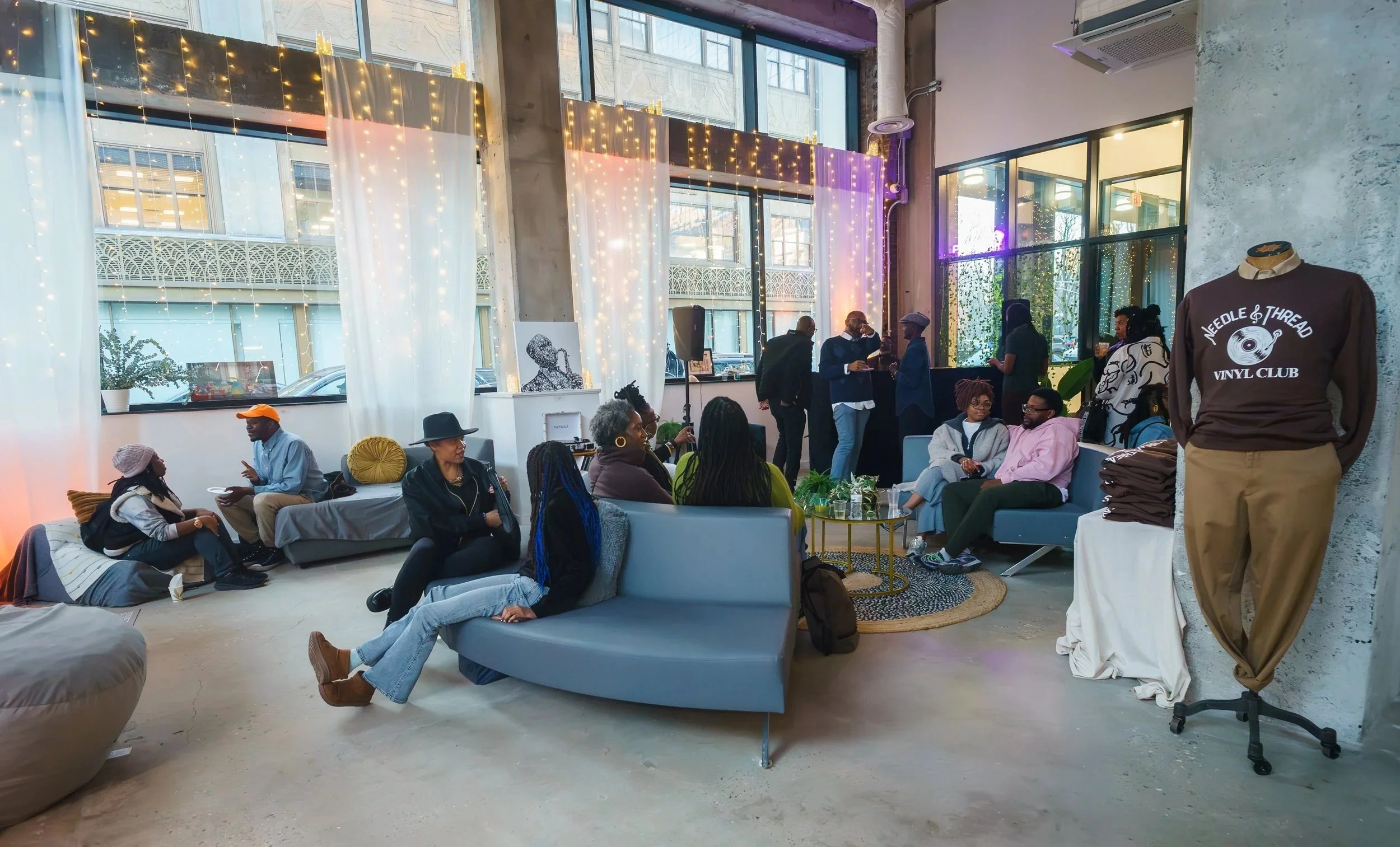 People socializing and sitting on couches in a decorated indoor setting with large windows, string lights, and art displayed on the walls, resembling a social or community event.