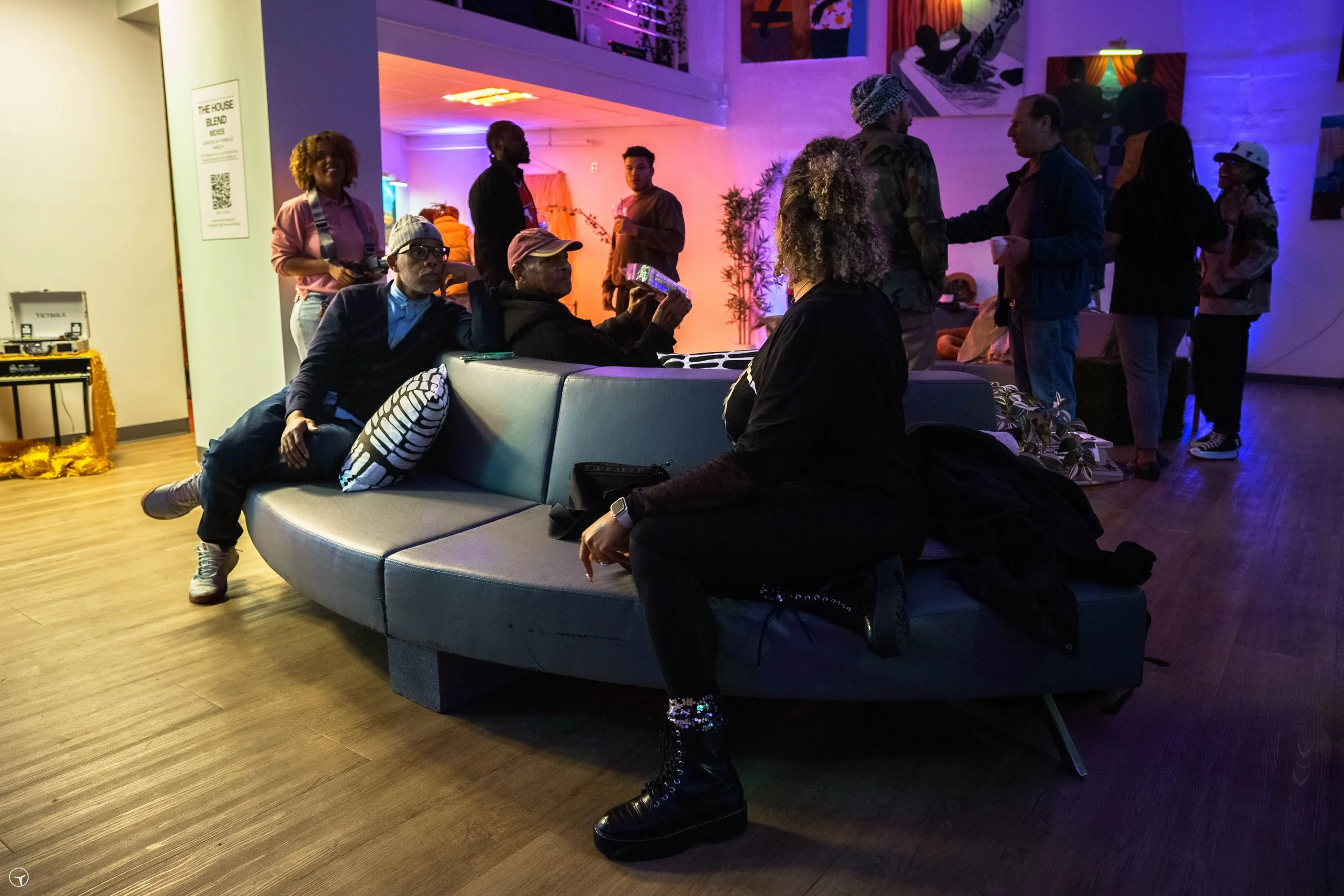 A group of people socializing in a dimly lit, colorful lounge or art gallery space, with some seated on a modern curved sofa and others standing or walking in the background.