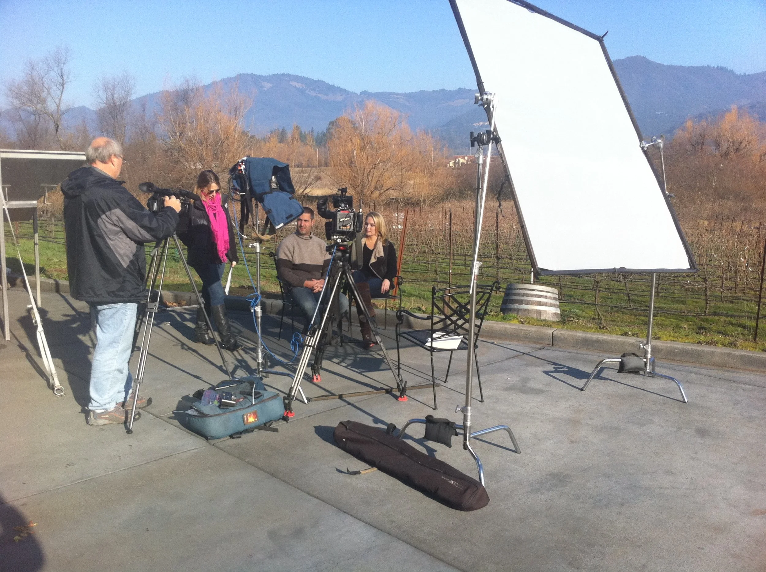 Filming setup outdoors with two people sitting in chairs in front of a camera, crew members operating cameras, lights, and reflectors, with a scenic mountain landscape in the background.