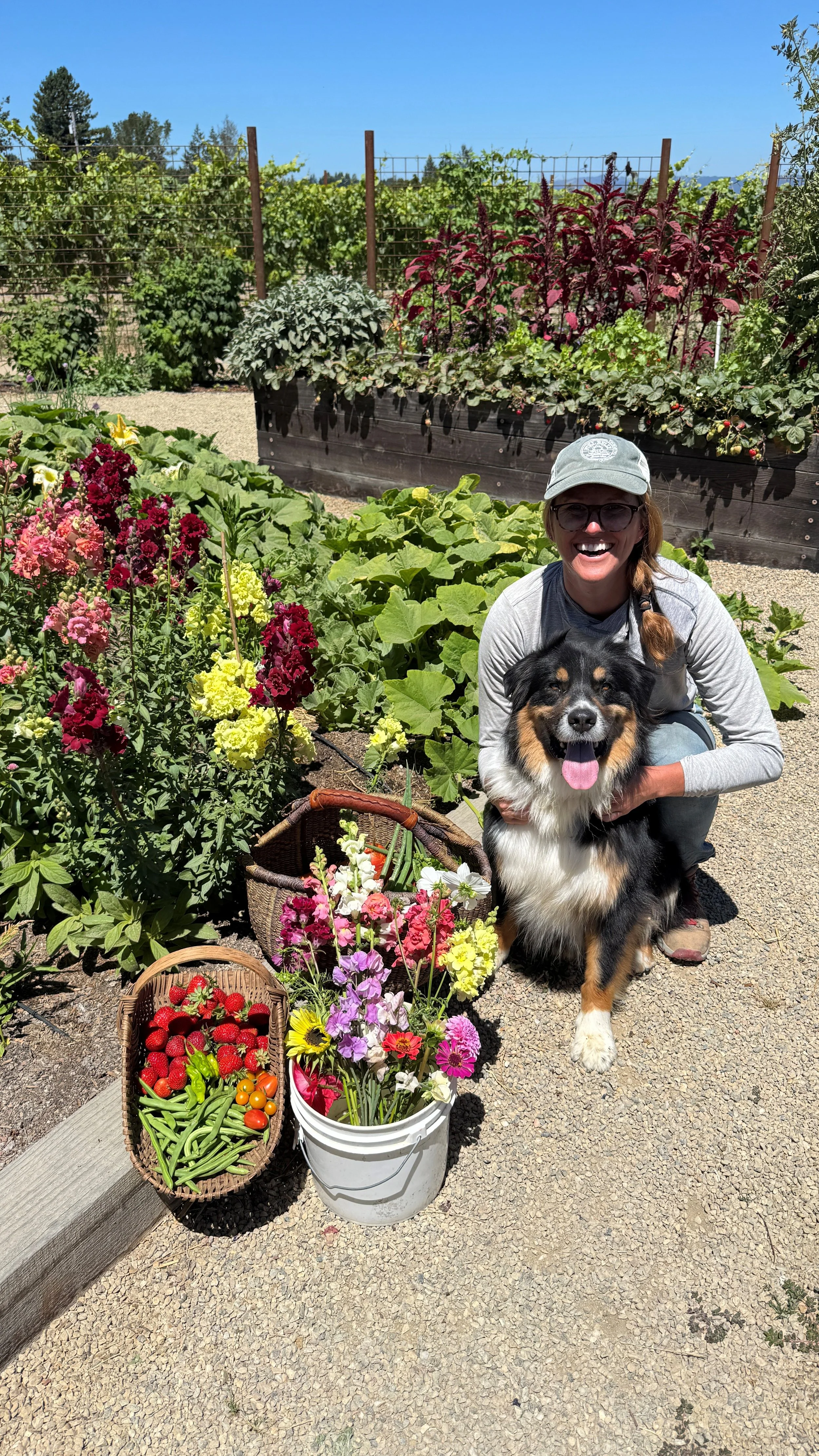 Woman in sunglasses smiling with a black and tan dog among flowers and vegetables in a garden on a sunny day.
