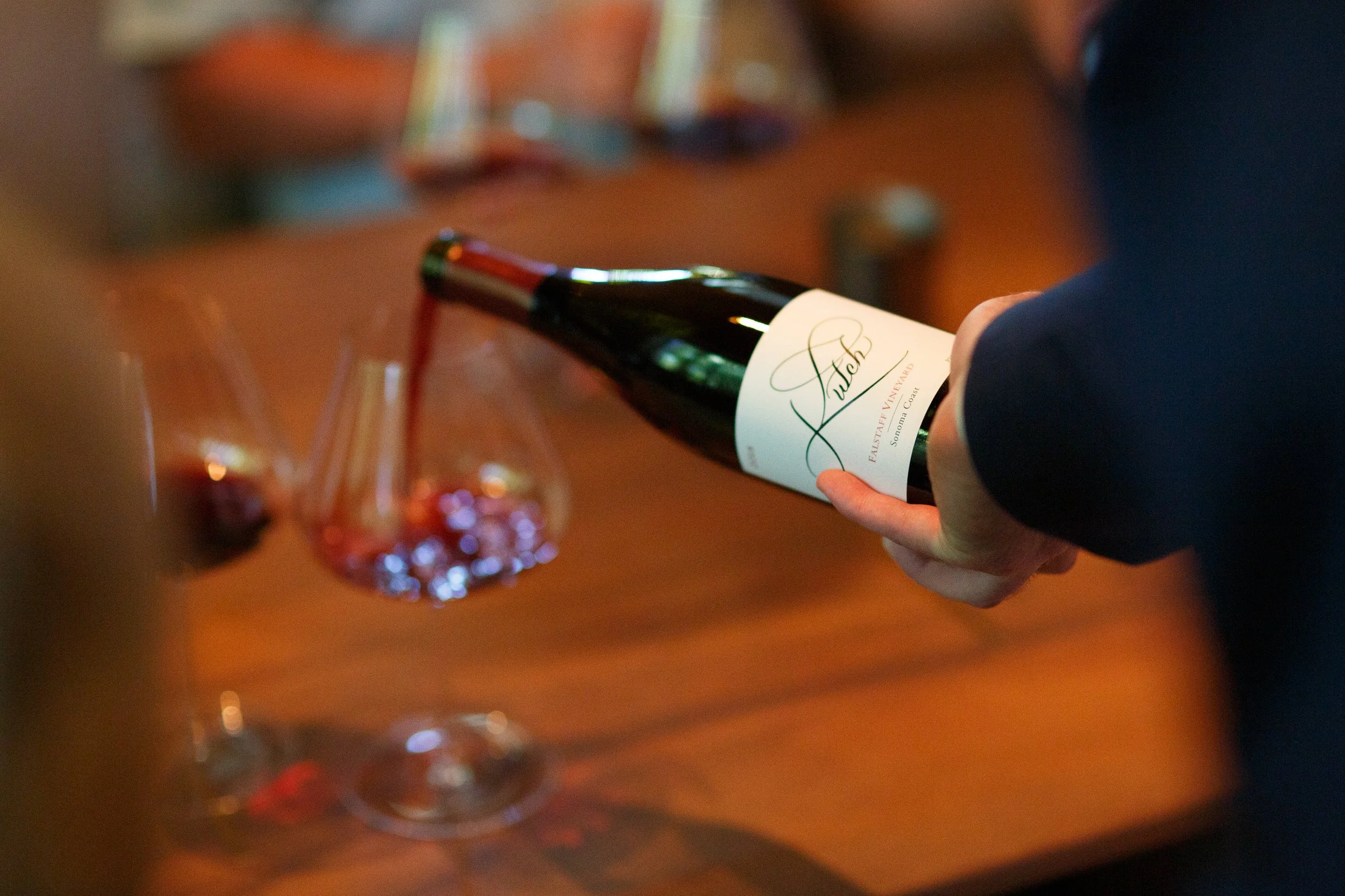 A person holding a bottle of Fallet Pas de Vigny sparkling wine over a wooden table with a glass of rosé wine in the foreground.