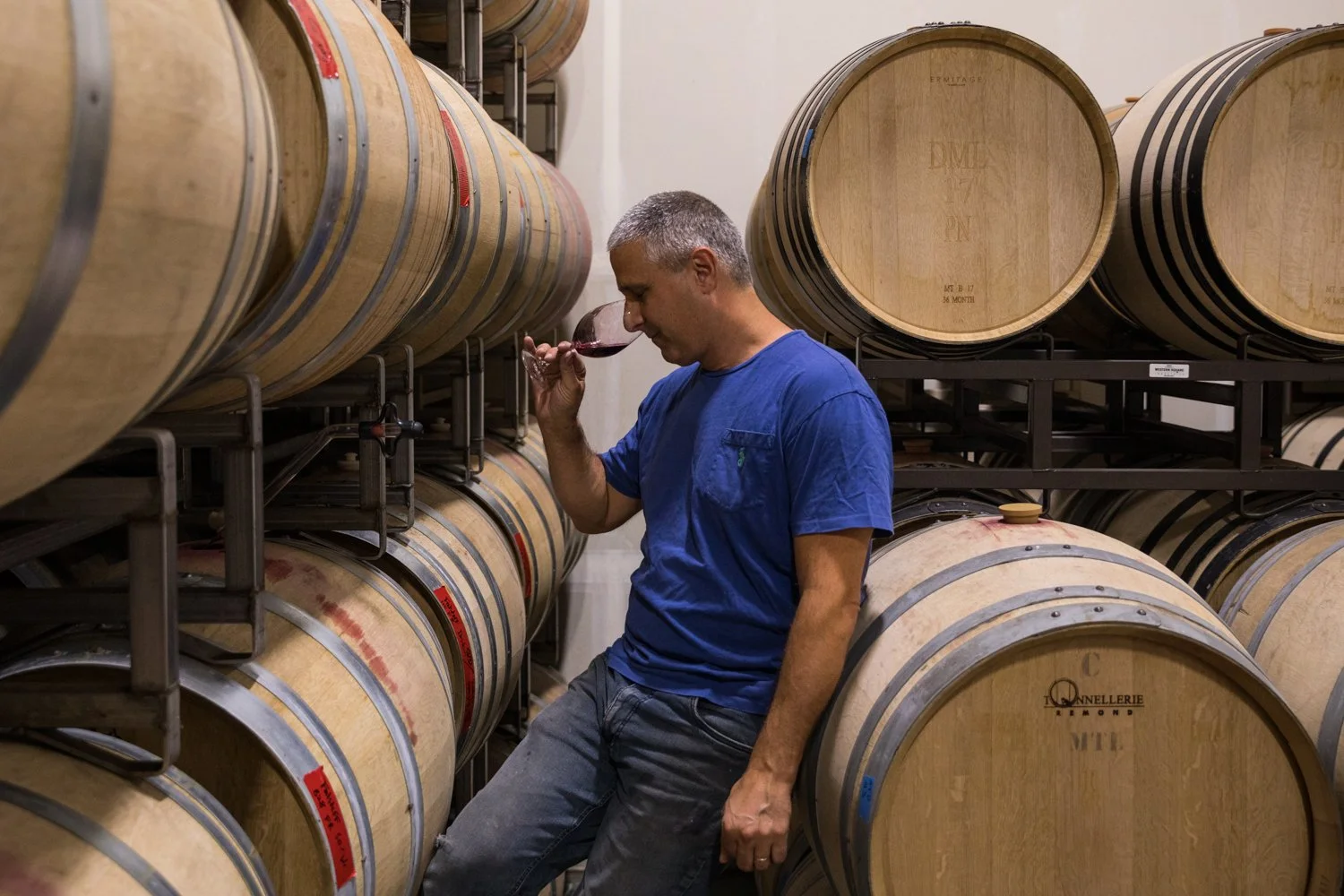 A man in a blue shirt tasting red wine in a wine cellar filled with wooden barrels.