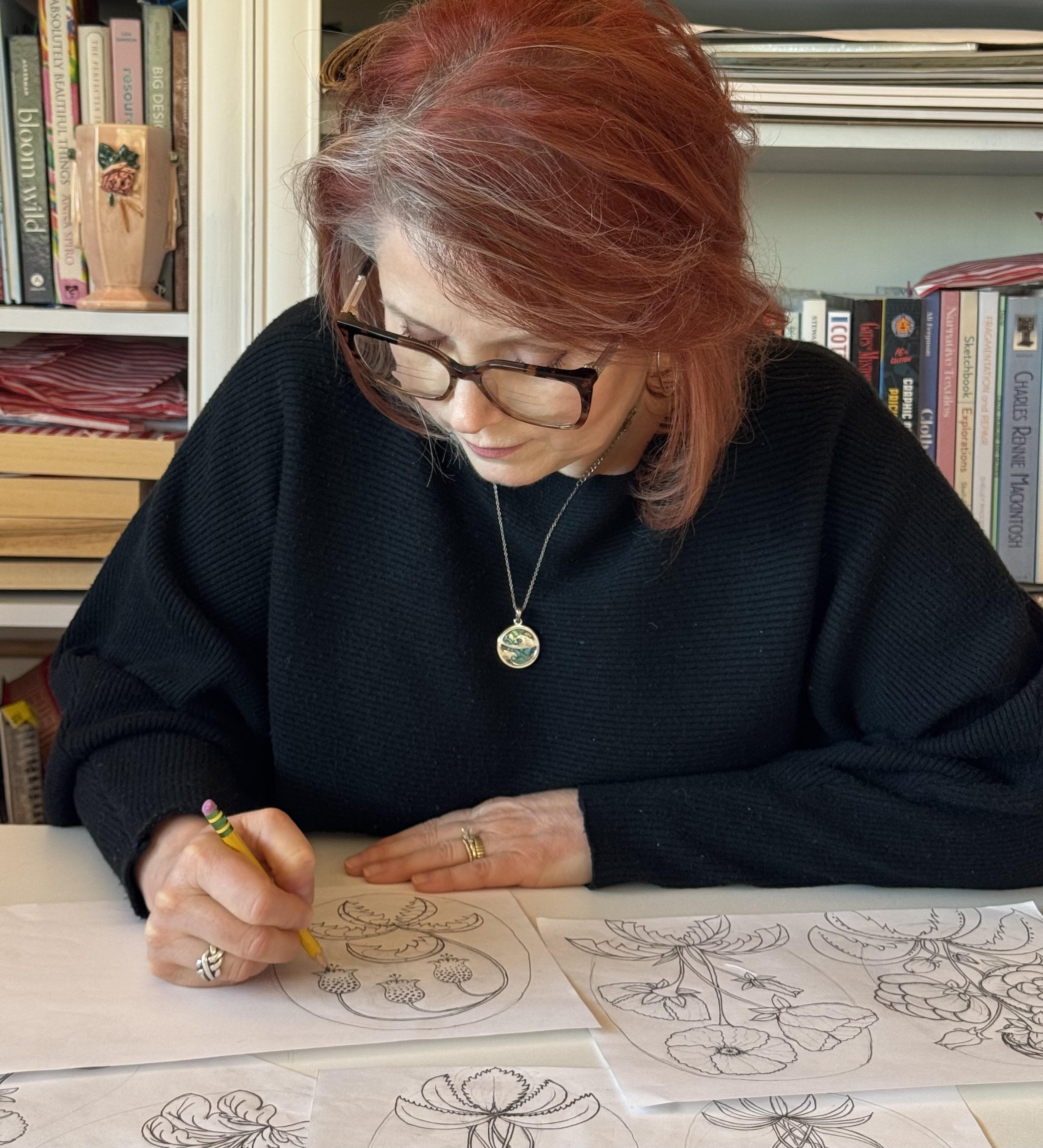 A woman with red hair and glasses is drawing floral patterns on paper with a yellow pencil. She is wearing a black sweater and a round pendant necklace. In the background, there are shelves filled with books and folded fabrics.