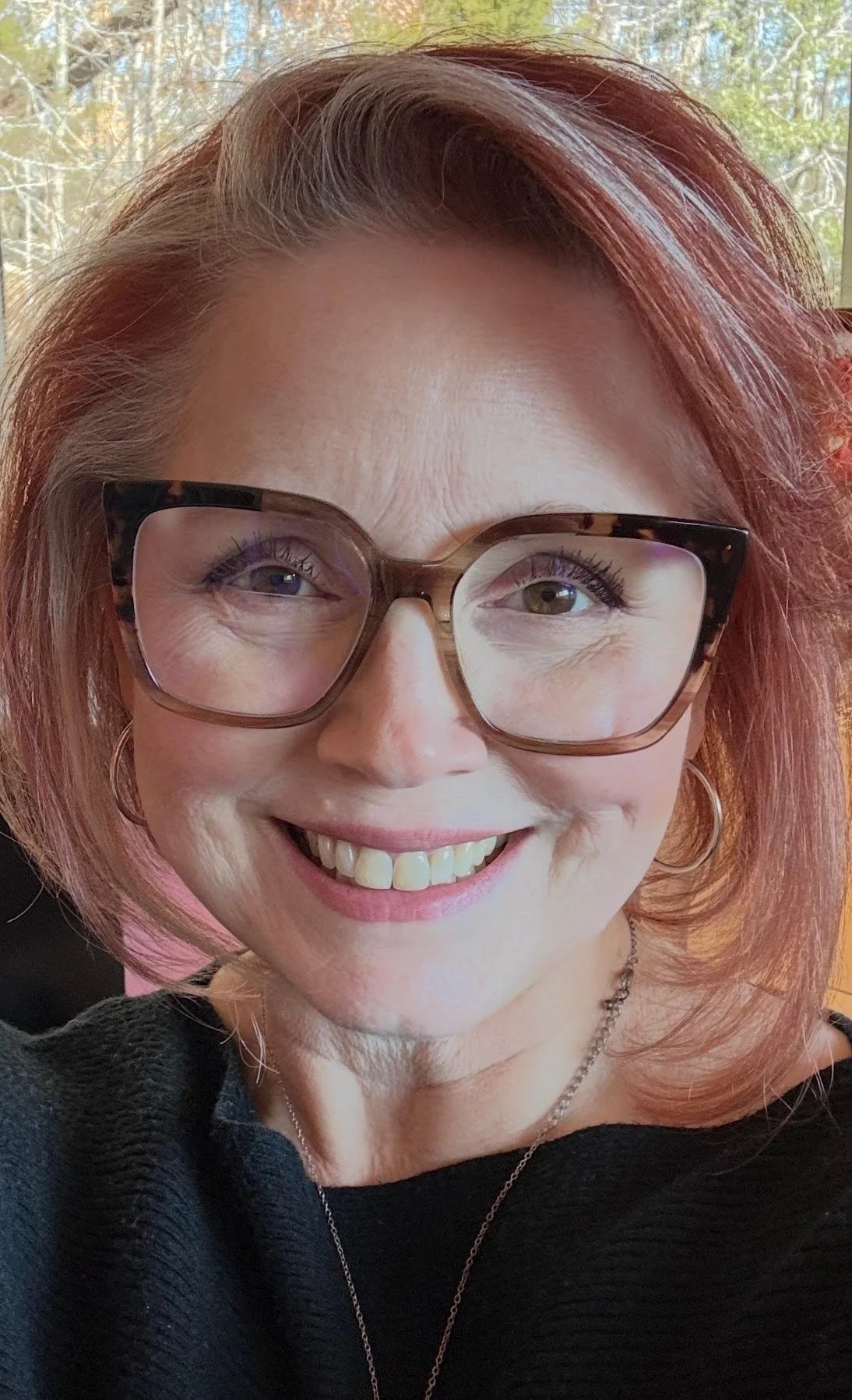 A woman with reddish hair, wearing large tortoiseshell glasses, smiling, with a background of trees and bright daylight.