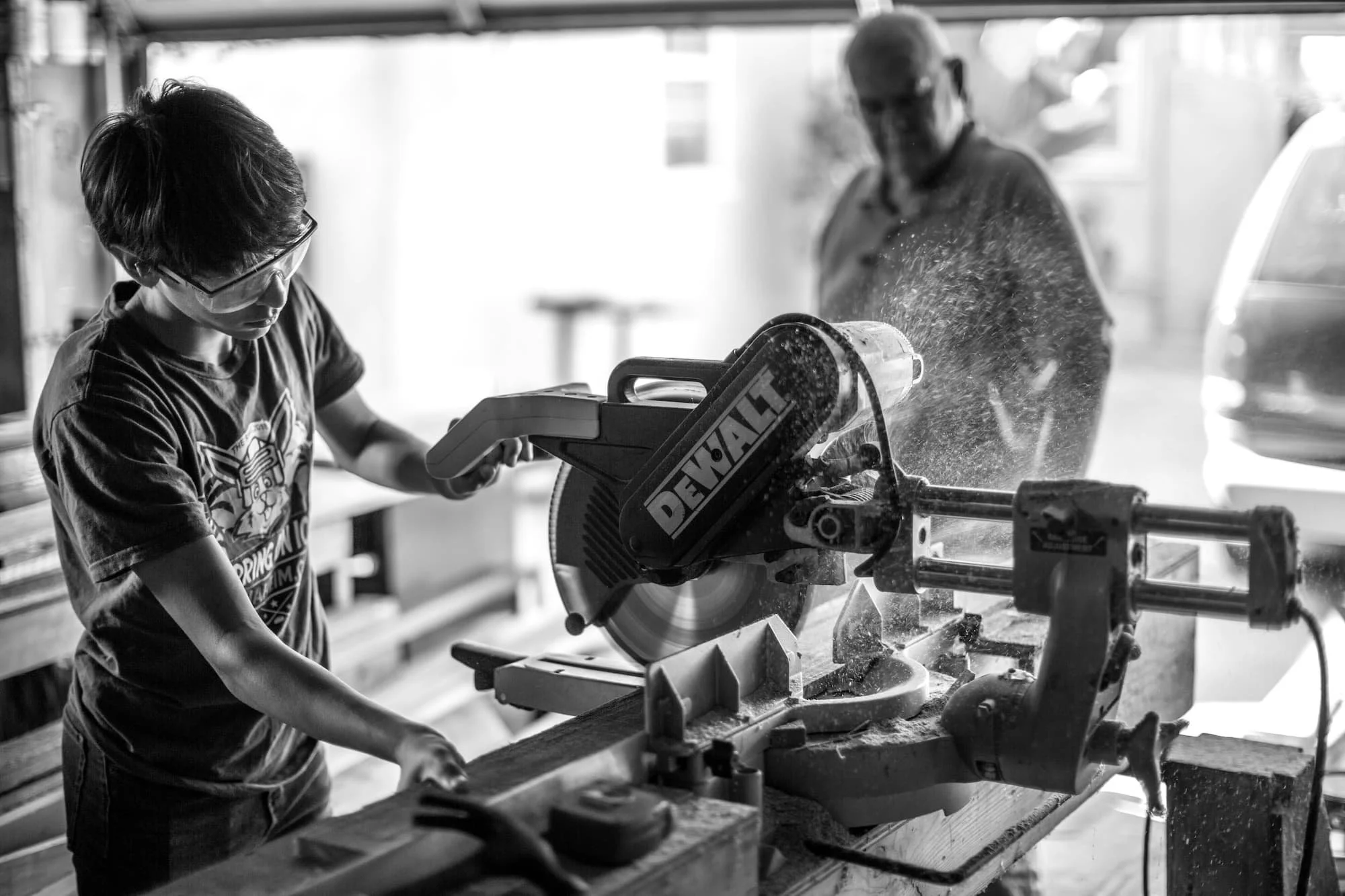 Young boy wearing glasses operating a DeWalt miter saw with an older man observing in a workshop.