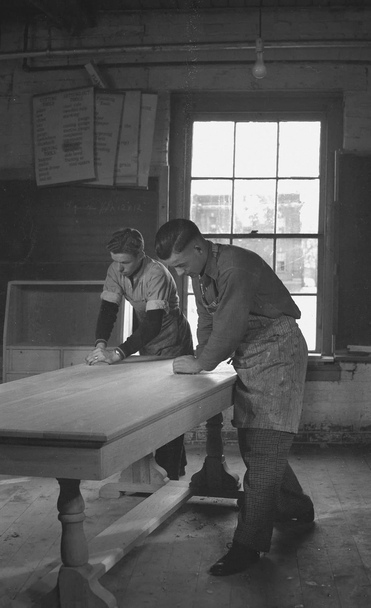 Two men are working on a wooden table inside a workshop with a large window. One man is sanding the table surface, and the other man is pressing down on the wood. There are signs or menus hanging on the wall above them.