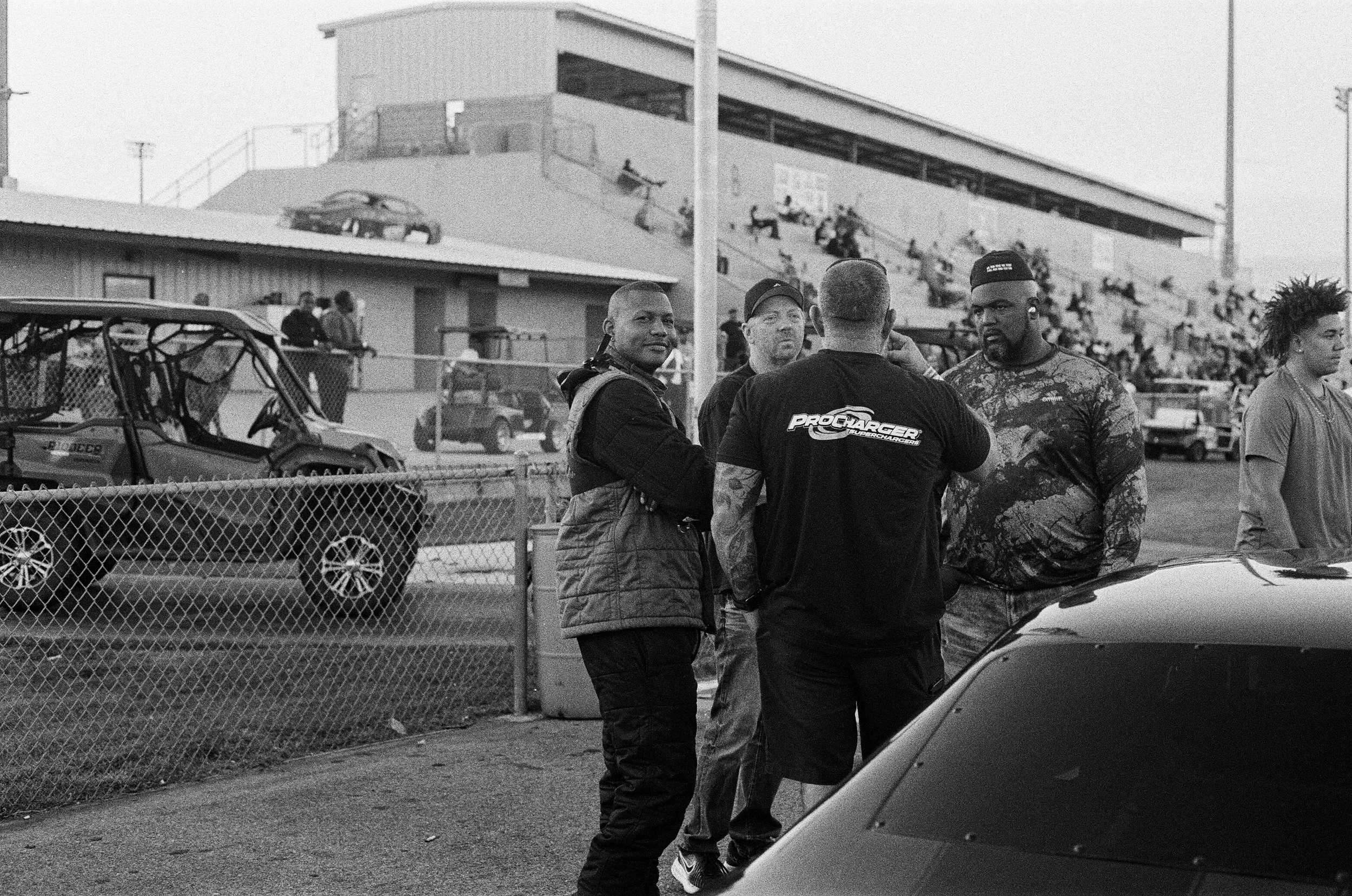 A group of people standing near a fence at a sports stadium, with a small vehicle and a building with bleachers in the background. It appears to be taken during daytime.