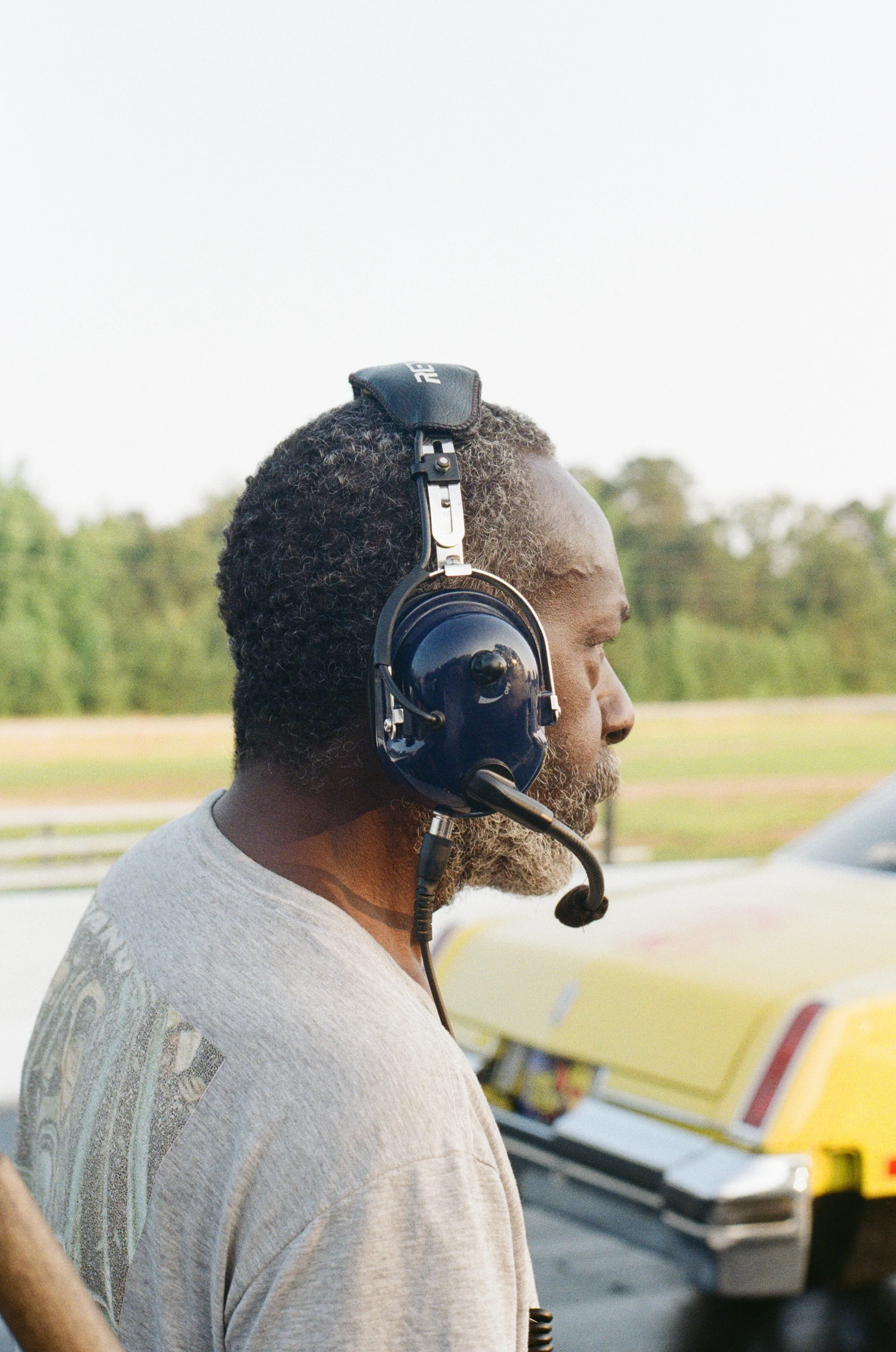 A man with dark, curly hair and a gray T-shirt wearing a headset with a microphone, standing outdoors with trees and a yellow vehicle in the background.