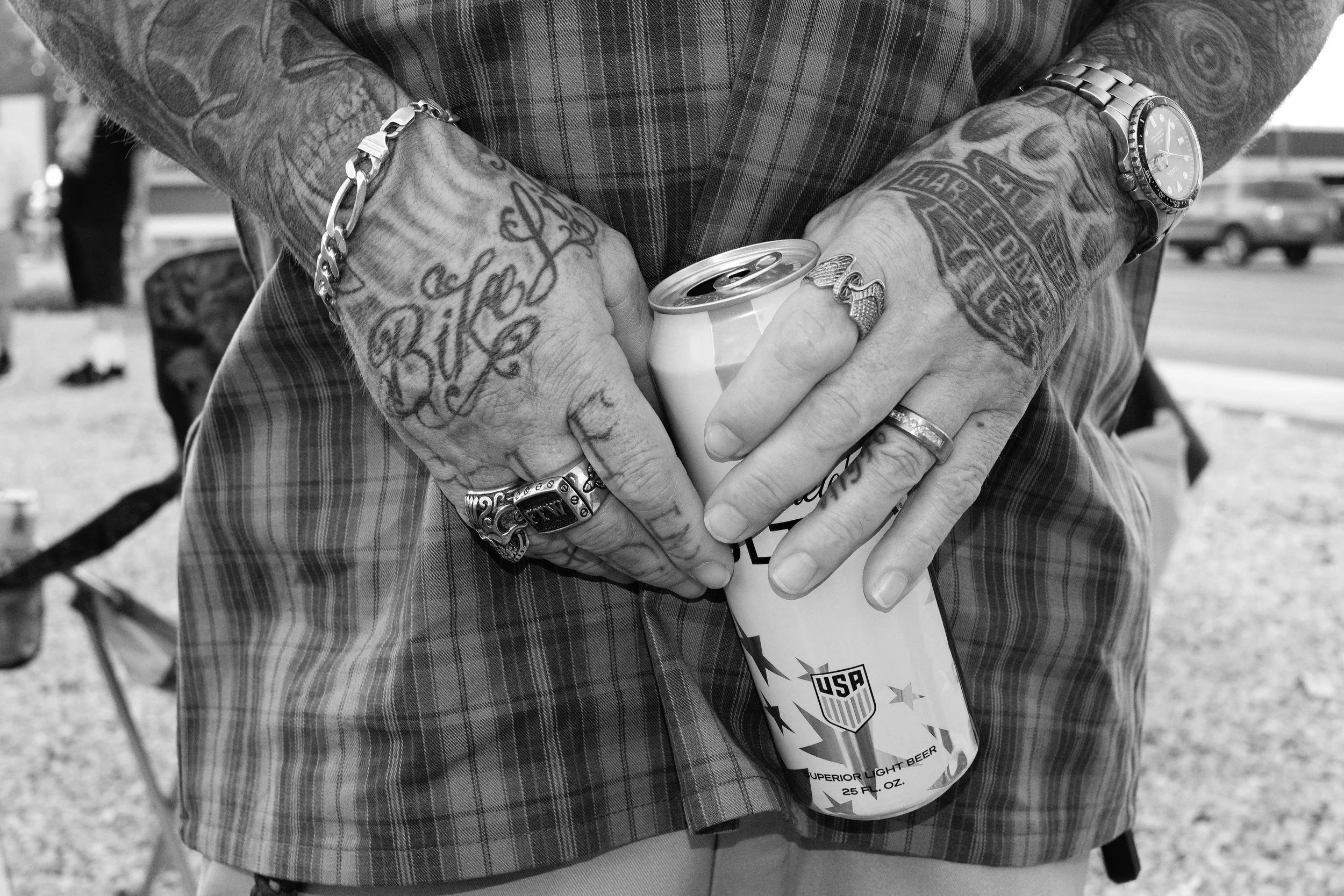 A person with tattoos on their arms, wearing a watch, bracelet, and rings, holding an open can of beer with a label that reads 'US' and 'super light beer' at an outdoor gathering.