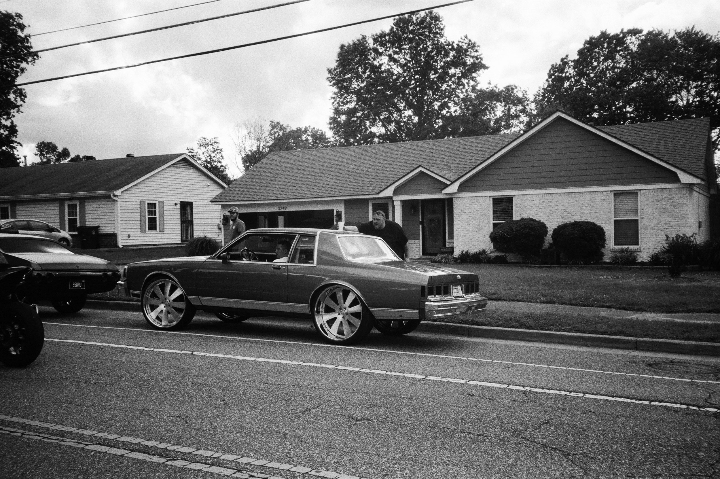 Black and white photo of a residential neighborhood street with parked cars, including a vintage car in the foreground and people walking on the sidewalk near houses with trees in the background.