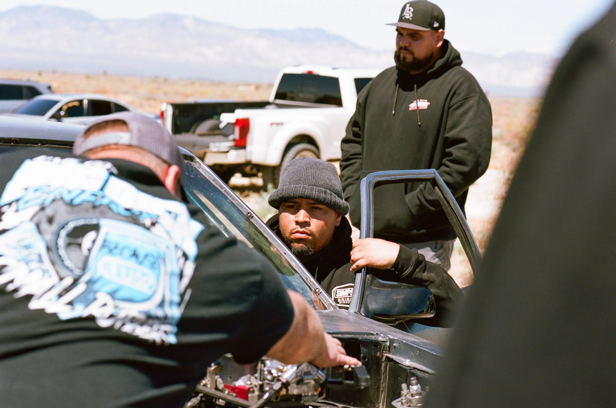 Several men working on a car outdoors in a desert-like area with mountains in the background. One man wearing a black hoodie and cap stands while others, one in a baseball cap, work on the car's engine.