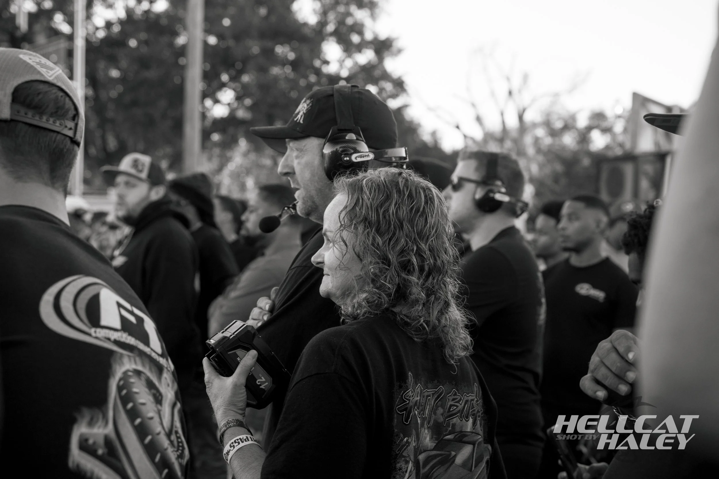 A group of people at a racing event, with one person in the center wearing a headset and holding a device, others wearing racing gear and hats, outdoors with trees in the background.