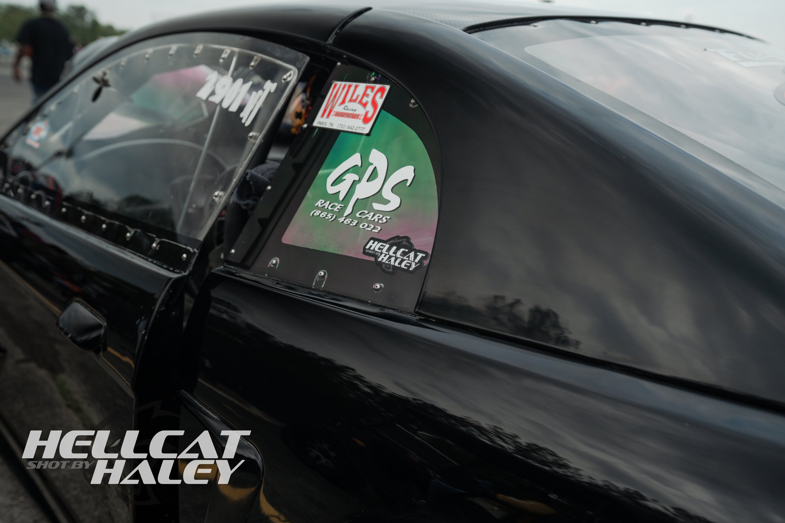 Close-up of a race car's side with stickers, including 'GPRS Race Cars,' a Hellcat sticker, and a sticker for 'Miless.' The car is black with a sleek design, and the photograph is taken outdoors with other people in the background.
