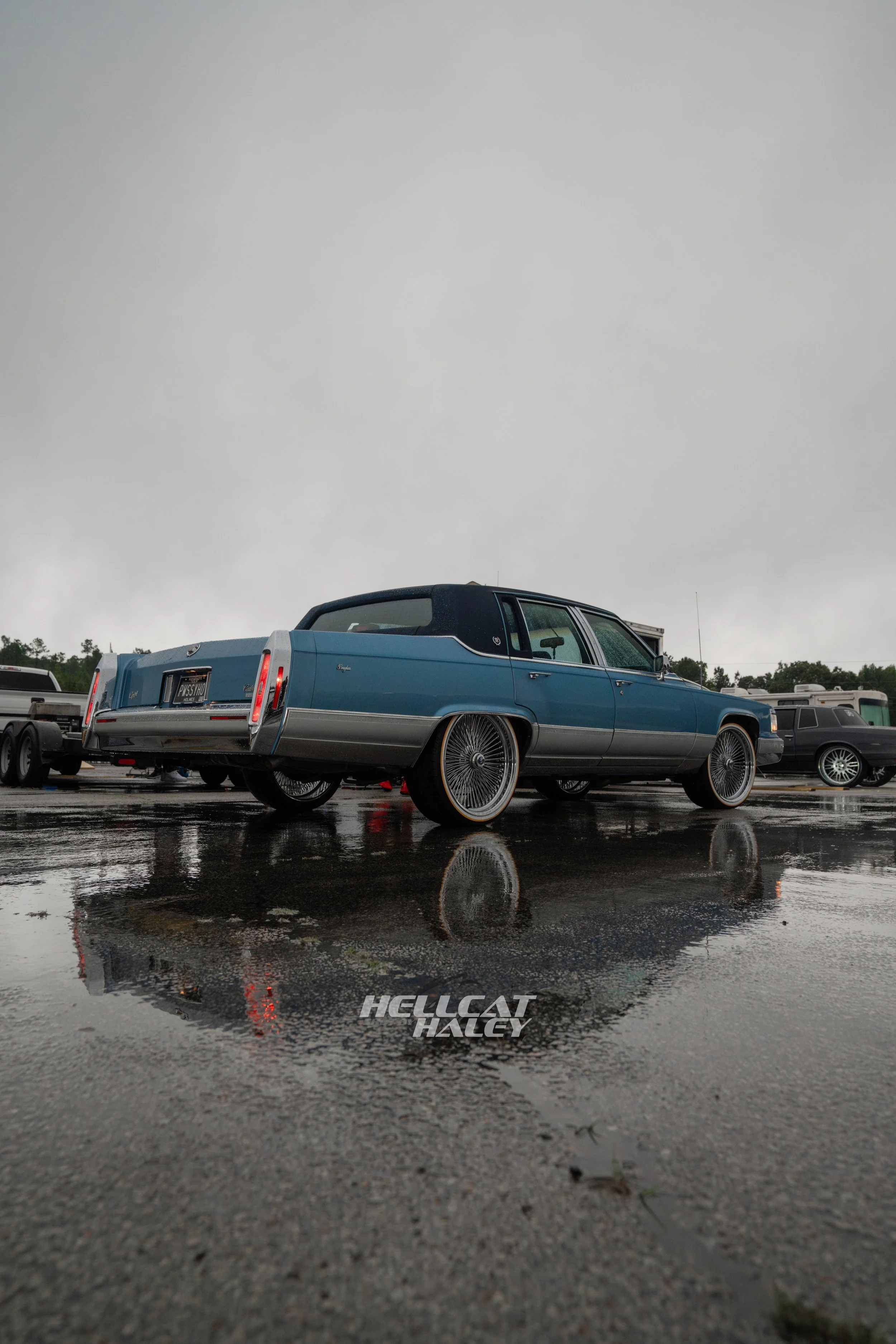 A vintage blue Cadillac parked on a wet parking lot with reflections of the car and the logo 'Hellcat Halcy' on the ground, under an overcast sky.