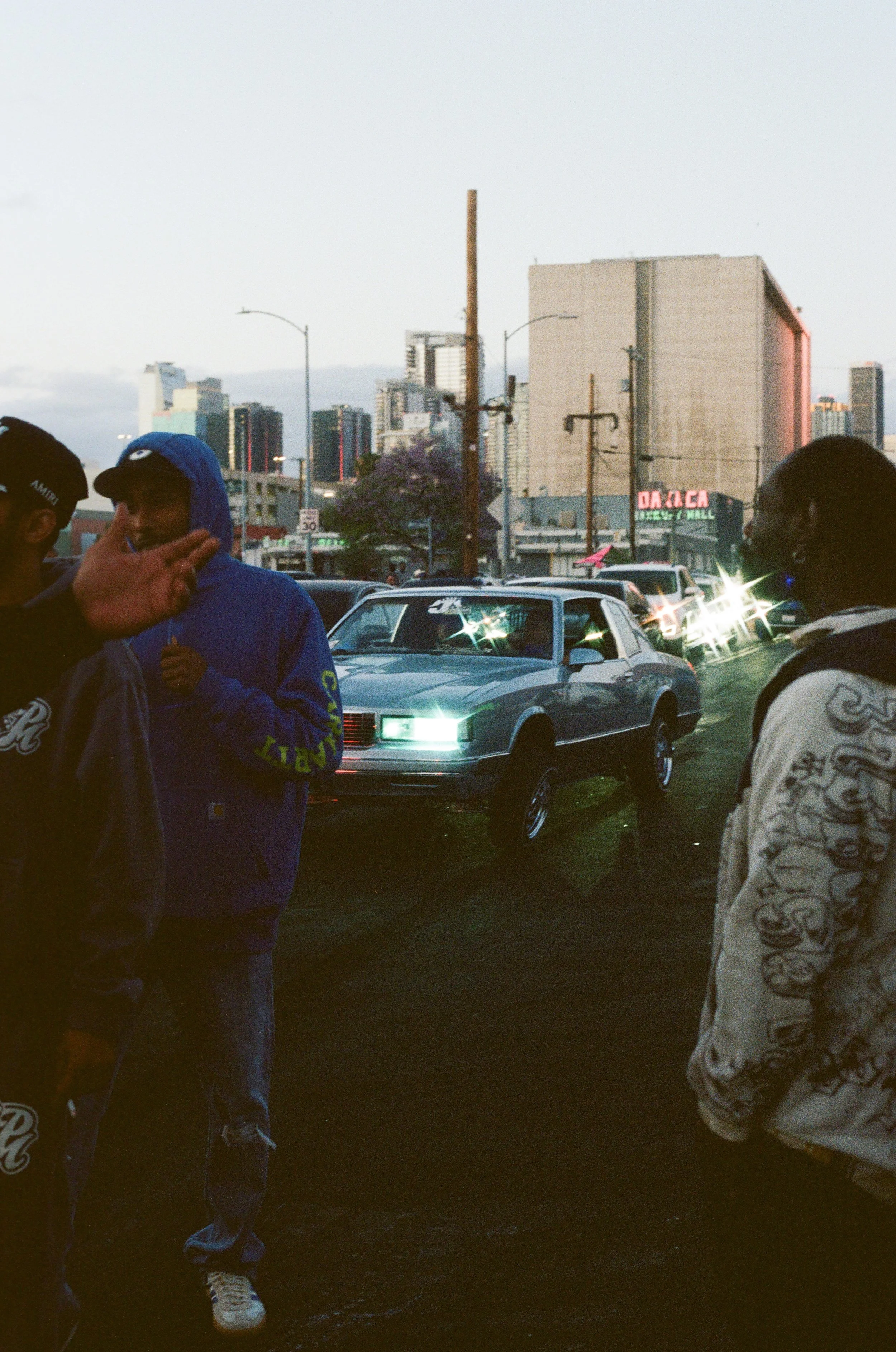 People standing on a city street with cars, tall buildings, and a sign reading 'Daiso' in the background, during dusk or early evening.