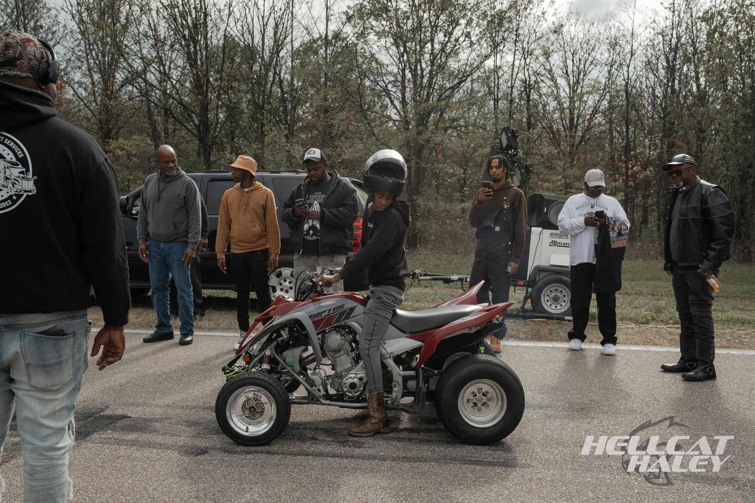 Group of people gathered outdoors around a red motorcycle, with trees in the background, some looking at their phones, and a person working on the motorcycle.