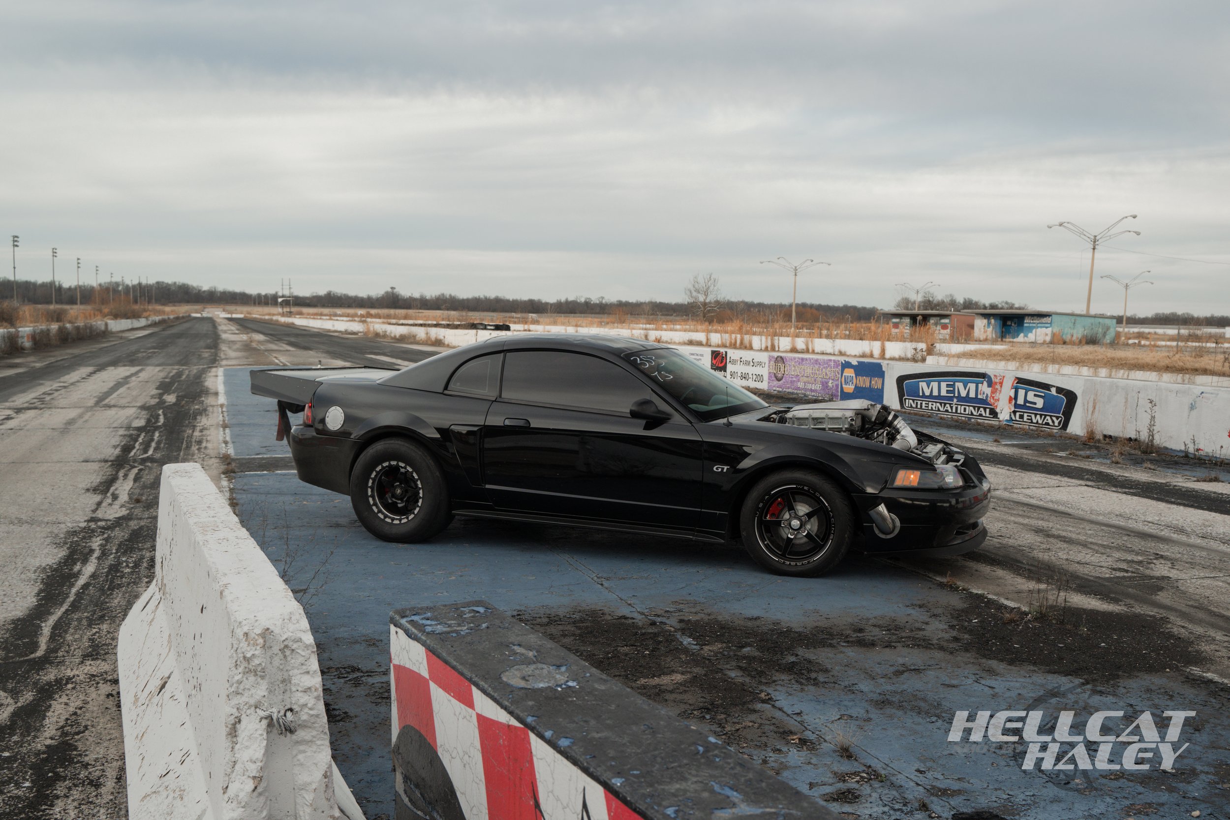 A black sports car with a damaged hood on a racetrack, with worn asphalt and barriers, under a cloudy sky.