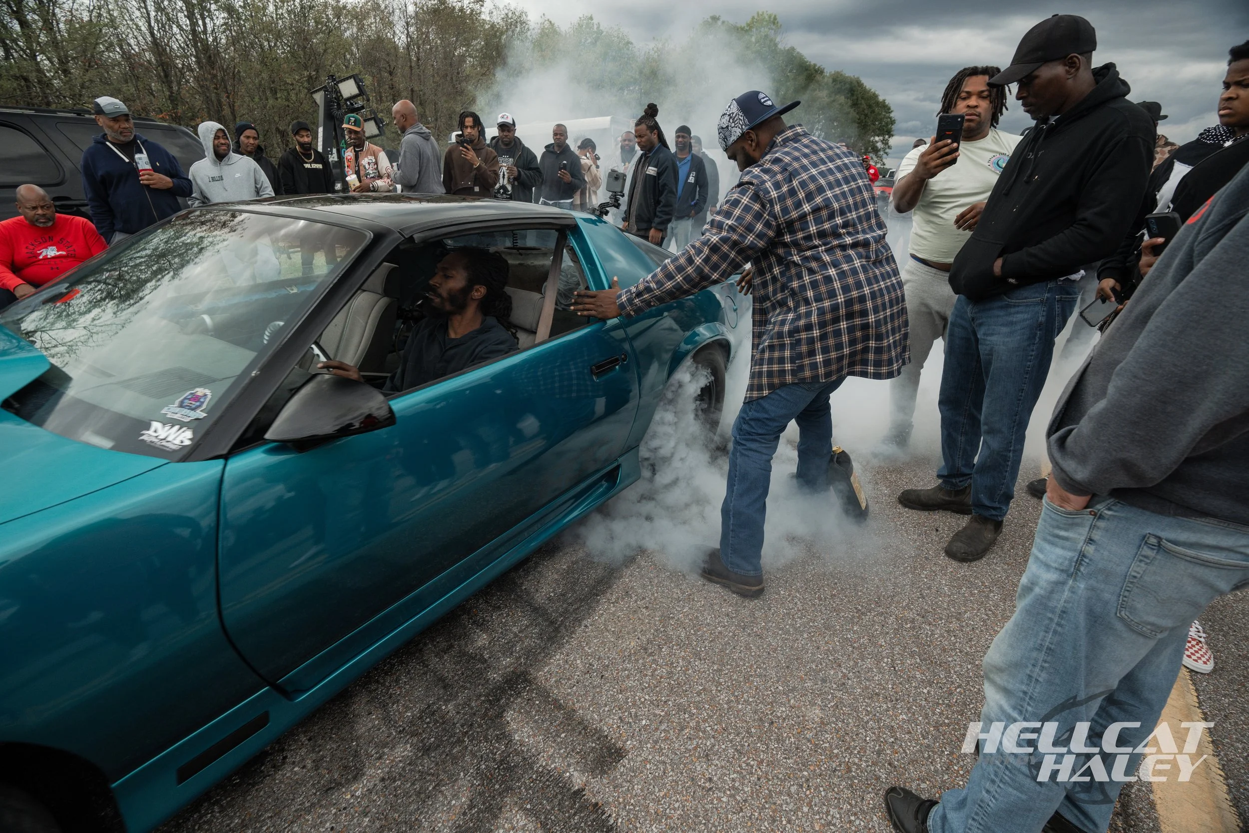 Group of people gathered at a car event, with a man pushing smoke out of a sports car while others observe and use their phones during overcast weather.