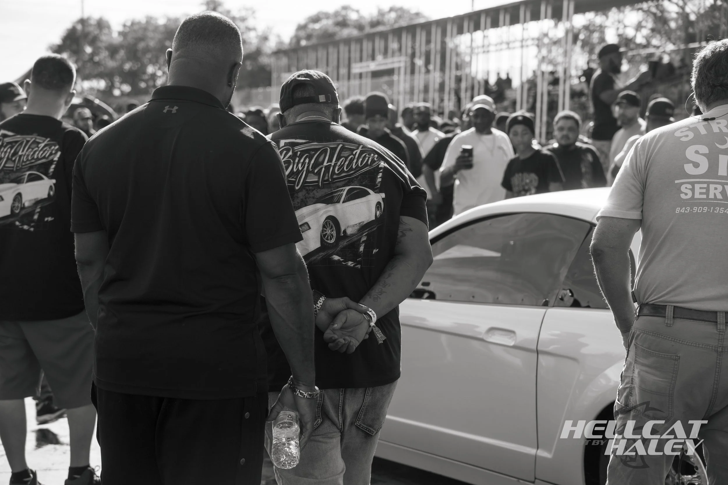 A group of people gathered outdoors, some wearing racing-related clothing, near a white car, with a chain-link fence and trees in the background, in black and white.
