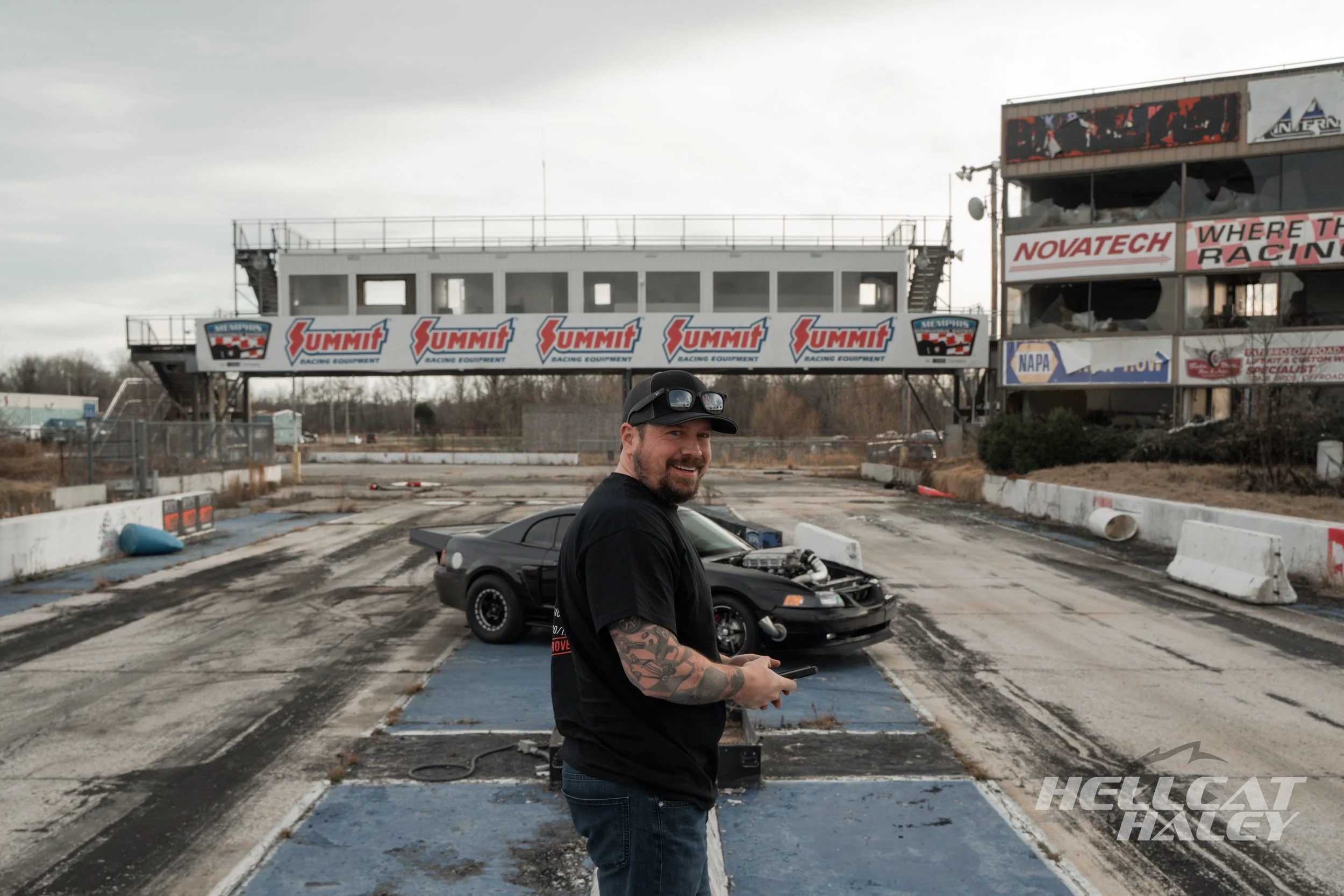 A man in a black t-shirt and jeans stands on a race track, holding a remote control, in front of a black sports car with its hood open. In the background, there is a large structure with multiple banners and advertisements, including Summit Racing Eq