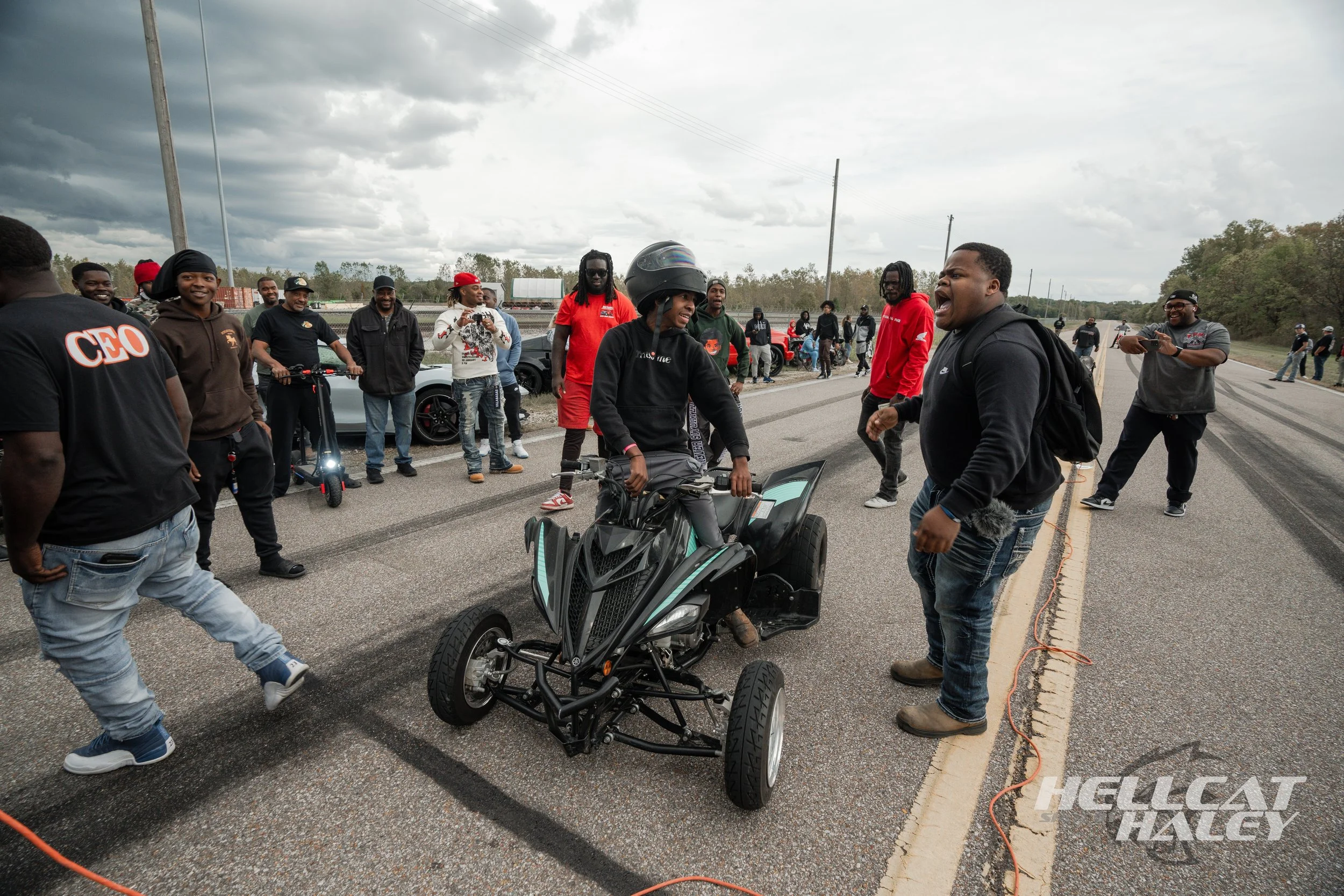 A group of people gathered on a road, with one person sitting on a black three-wheeled motorcycle, while others stand around, some smiling and talking. The sky is overcast, and there are cars and trees in the background.