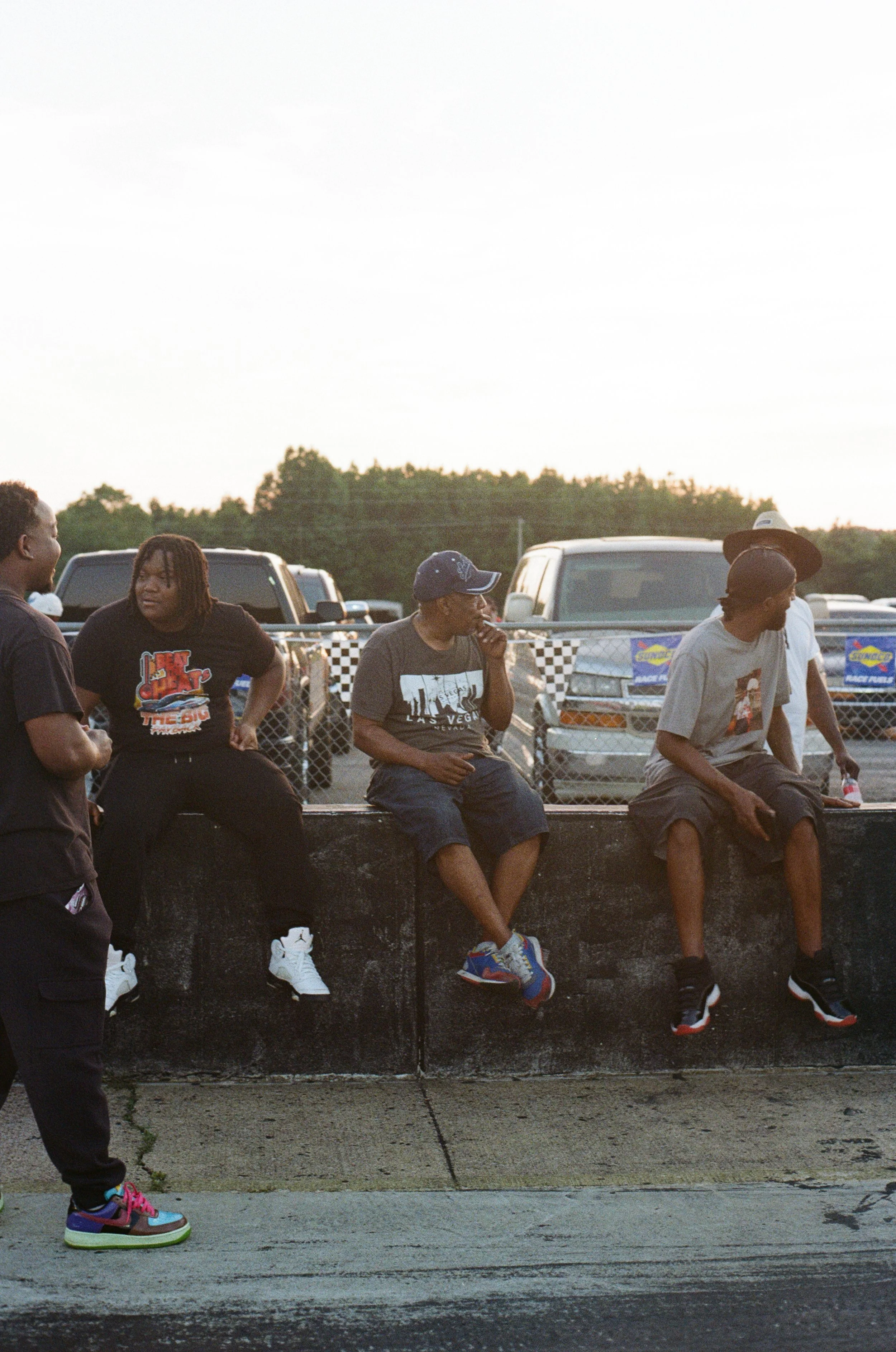 Four young men sitting on a black concrete ledge, with cars parked behind them and a chain-link fence with race banners, in an outdoor area during what appears to be late afternoon or evening.