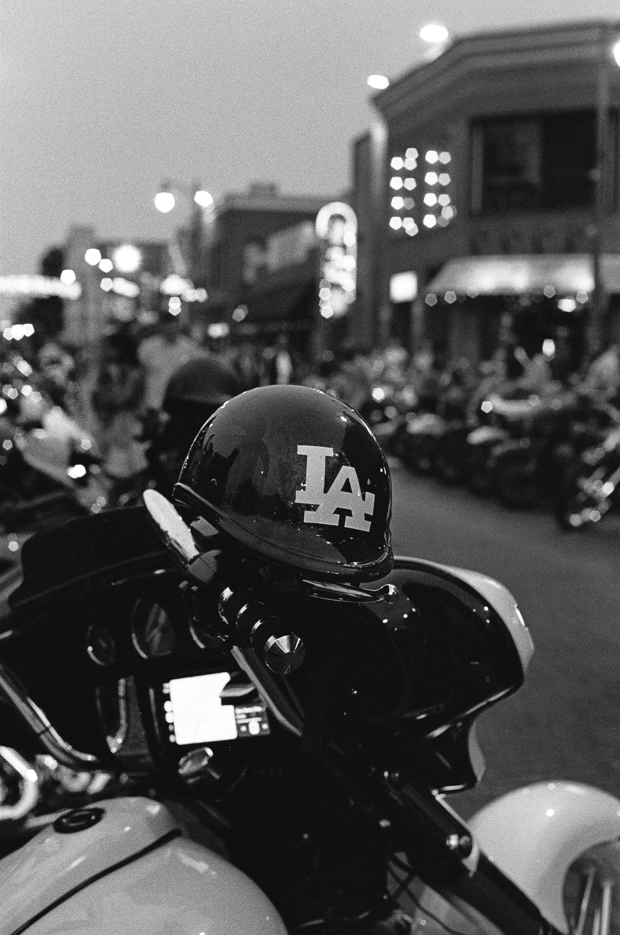 Black motorcycle helmet on a motorcycle with a Los Angeles Dodgers logo, parked among other motorcycles in an outdoor parking lot during evening or night.