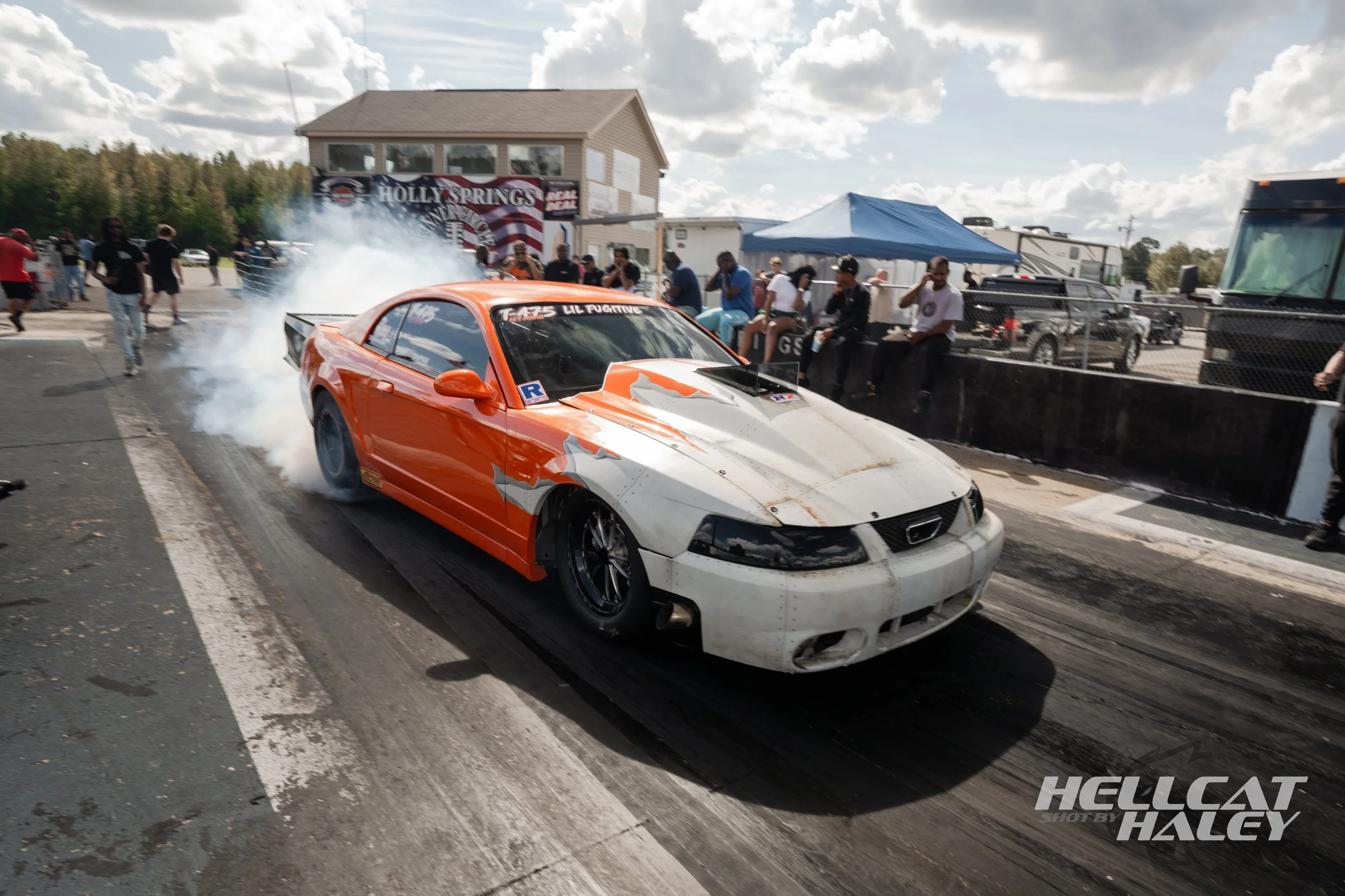A white and orange race car with damage on the front bumper emitting smoke at a race track with people watching and taking photos, a blue canopy tent, and a building with the sign "Holly Springs" in the background.