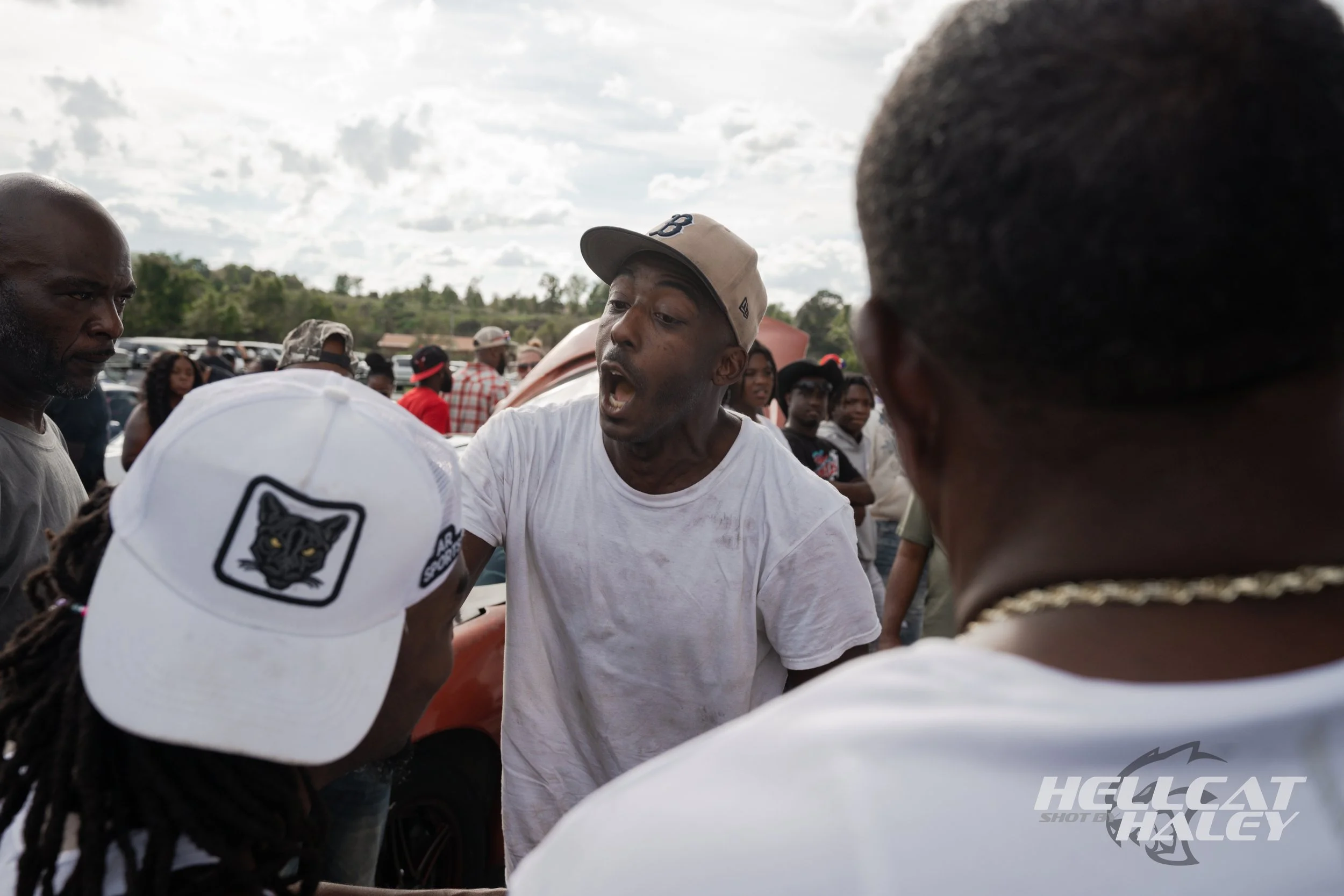 A man wearing a beige baseball cap and a white t-shirt is passionately speaking or arguing with others at an outdoor gathering. Several people are around him, some wearing white baseball caps with a black panther logo, and others are observing. The b