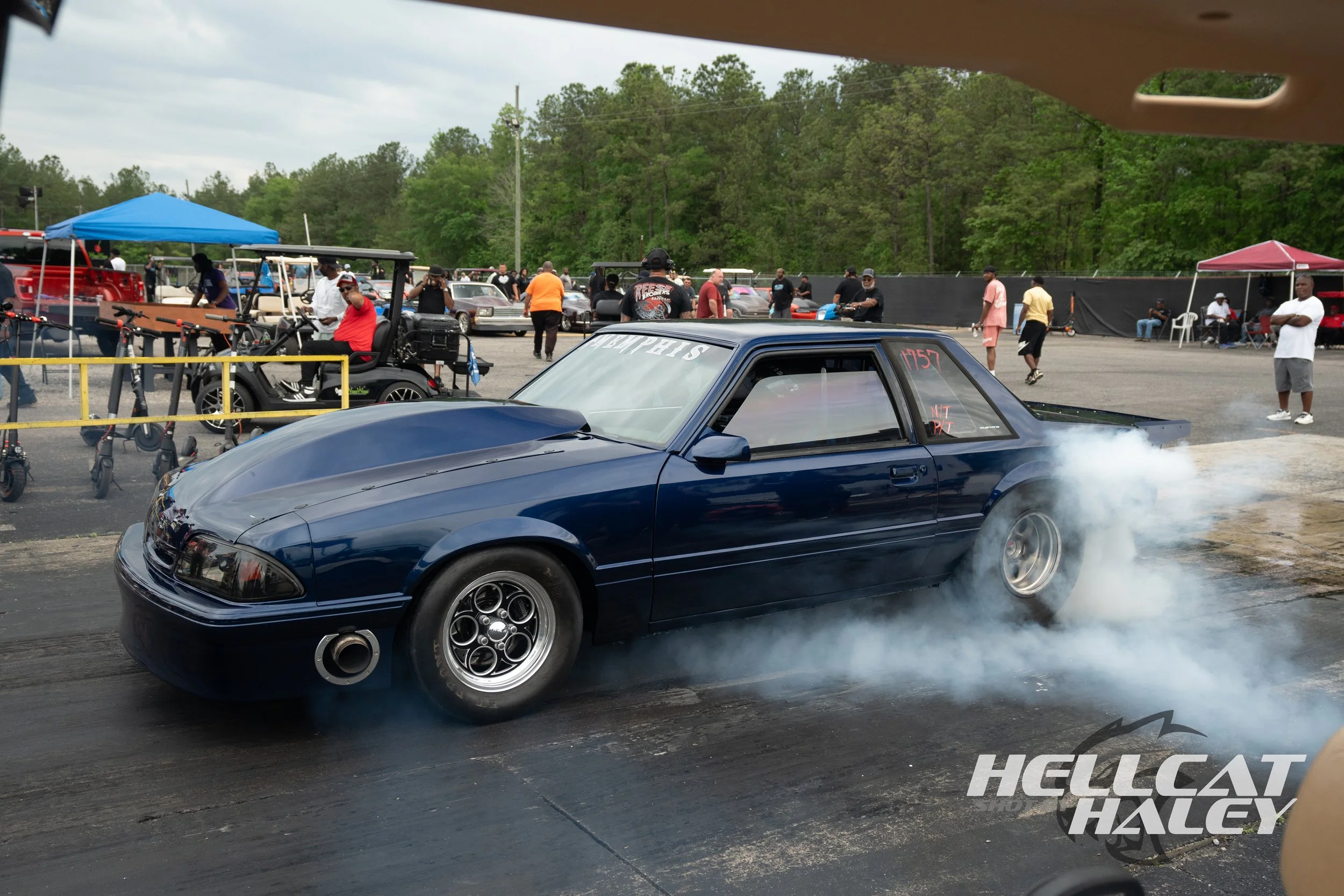 A blue sports car performing a burnout at a car event with smoke emanating from the tires, surrounded by people and other cars in the background.