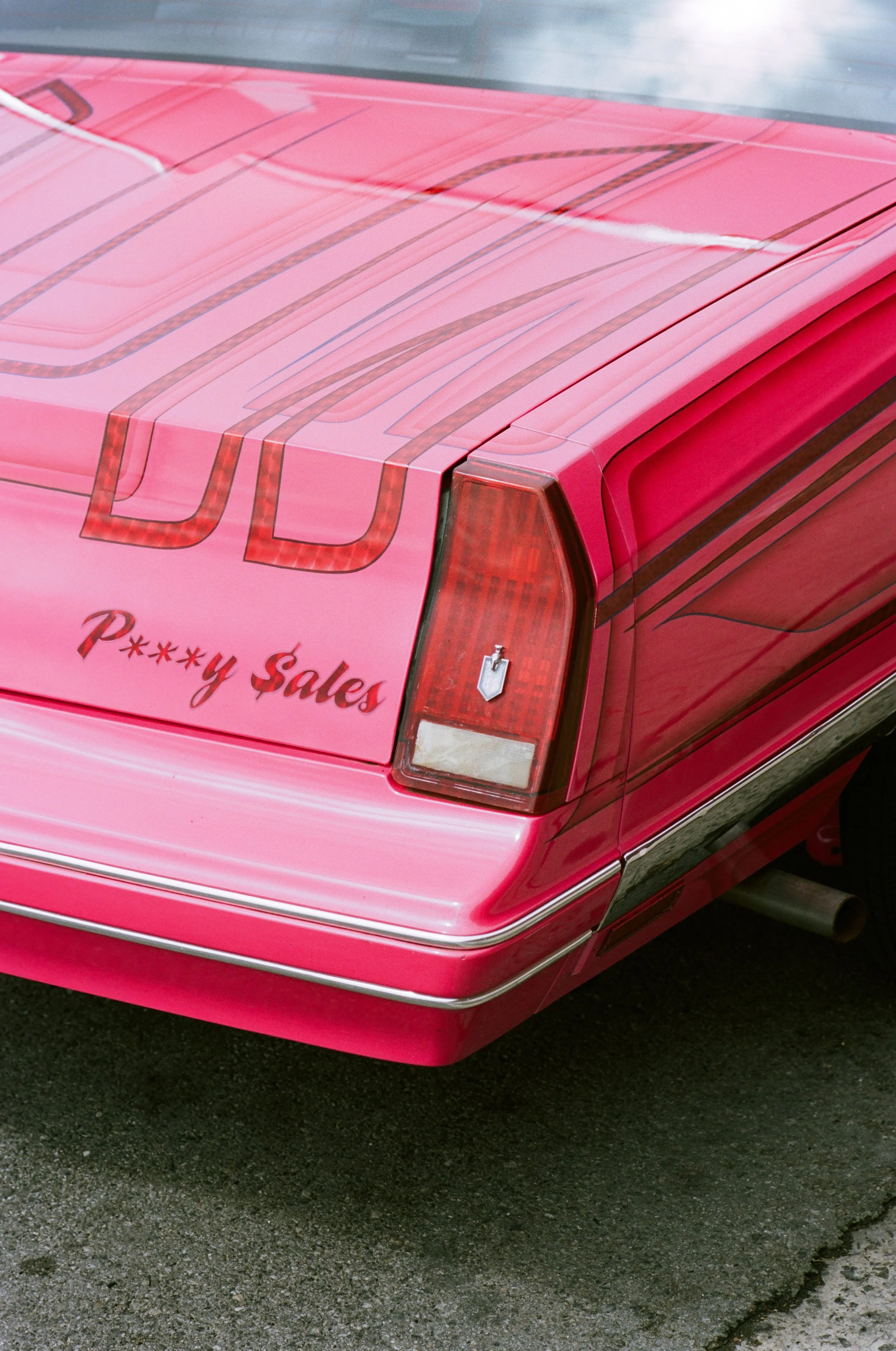 Close-up of the rear of a pink car with a scrolling decal that says 'P***y Sales' and a checkered stripe on the trunk and rear light.