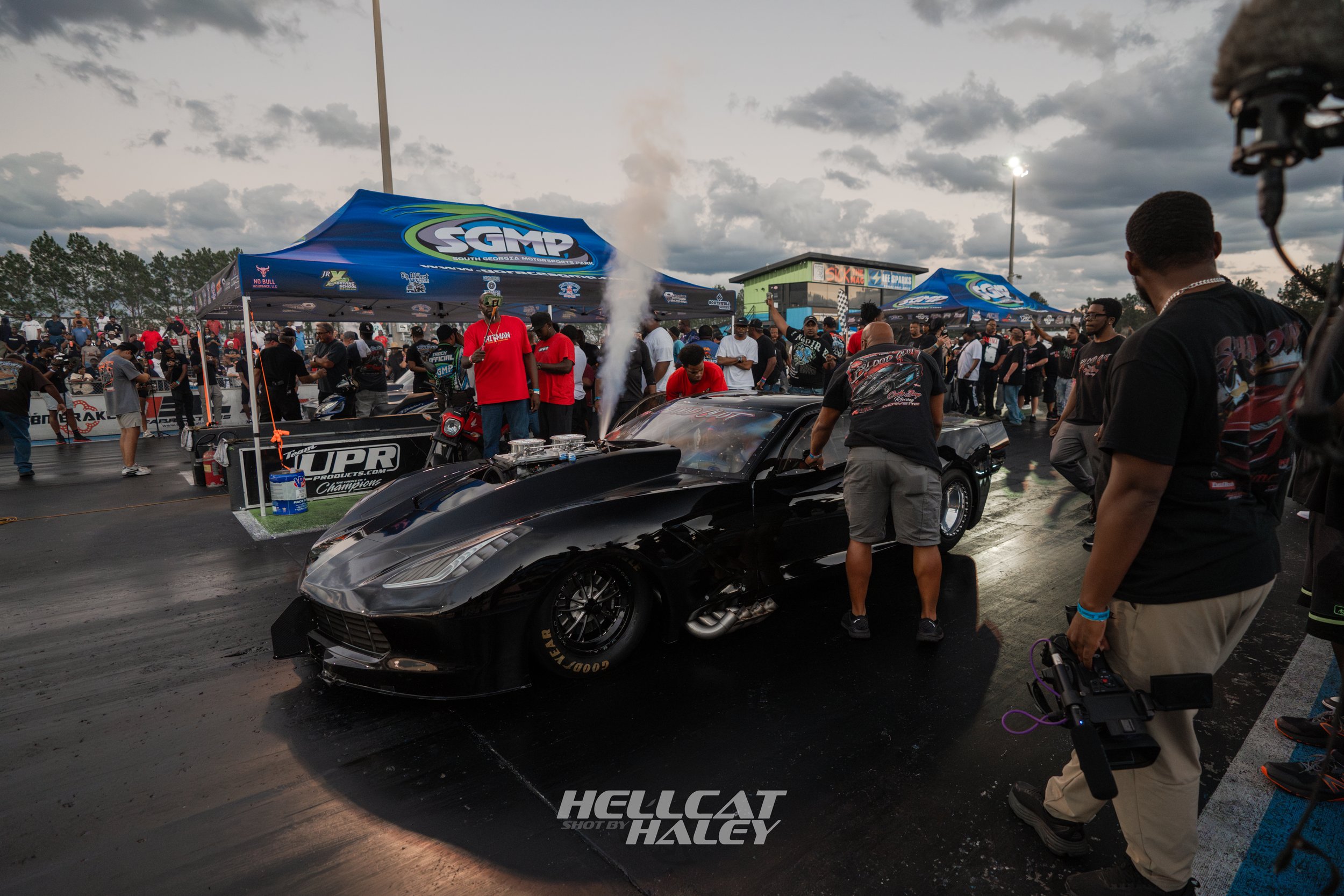 A drag racing event at South Georgia Motorsports Park with a black sports car on the track, people gathered around, and tents with race banners under a cloudy sky.