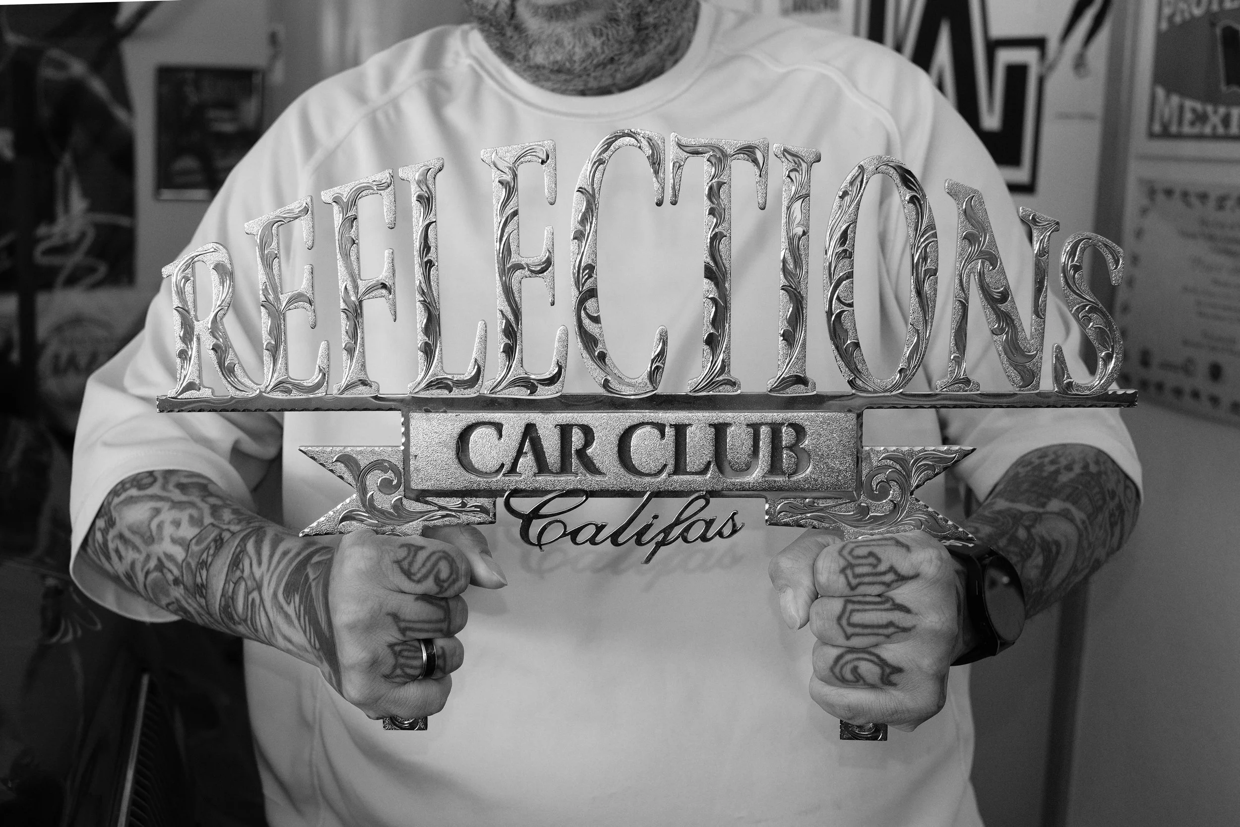 Person with tattoos on arms holding a large silver sign reading 'RELEASED' and 'Calipas' in a tattooed hand, in a room with posters and signs on the wall.